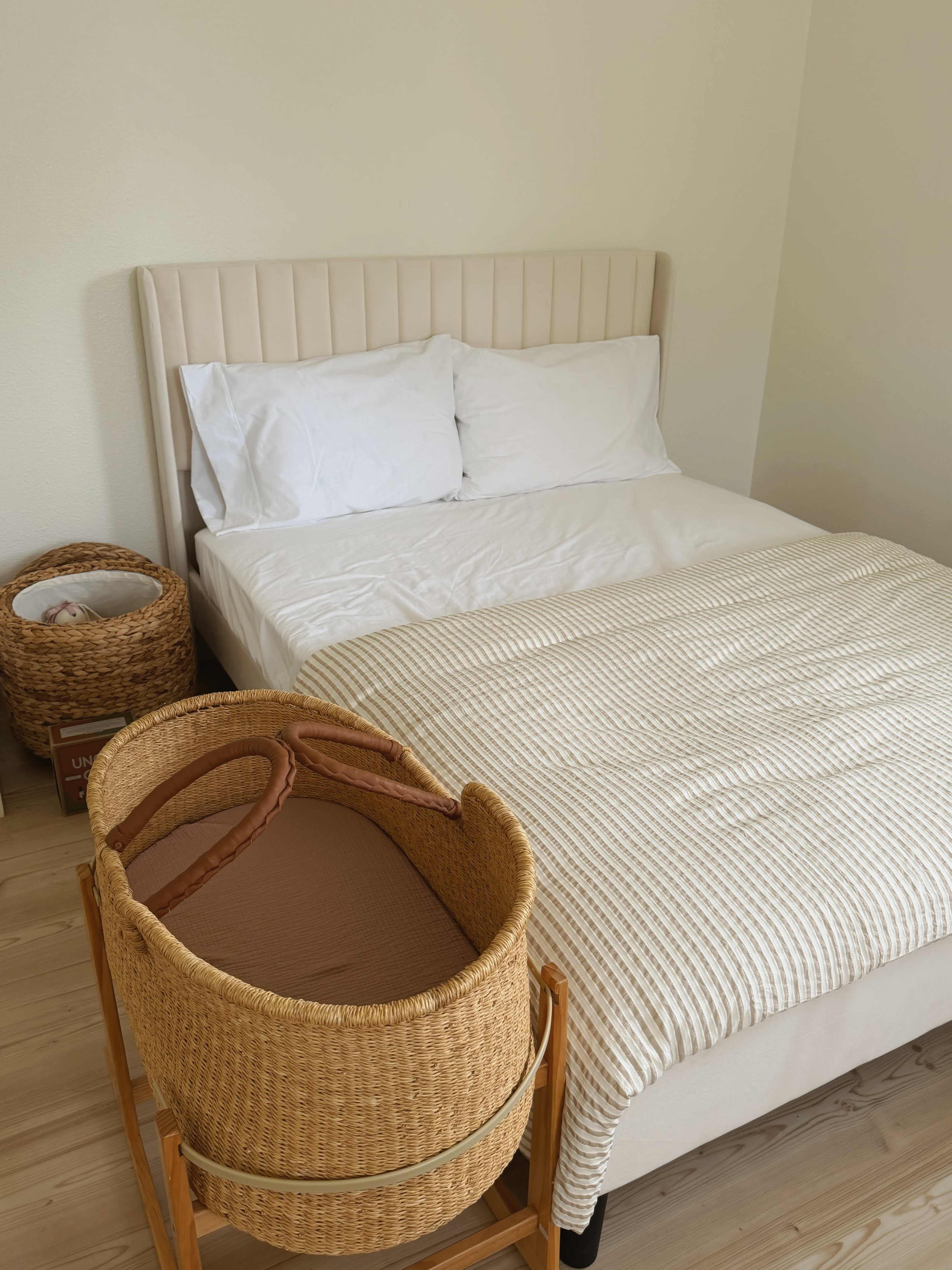 A light-colored bedroom features a neatly made bed with white sheets and two pillows, alongside a wicker bassinet on a wooden stand.