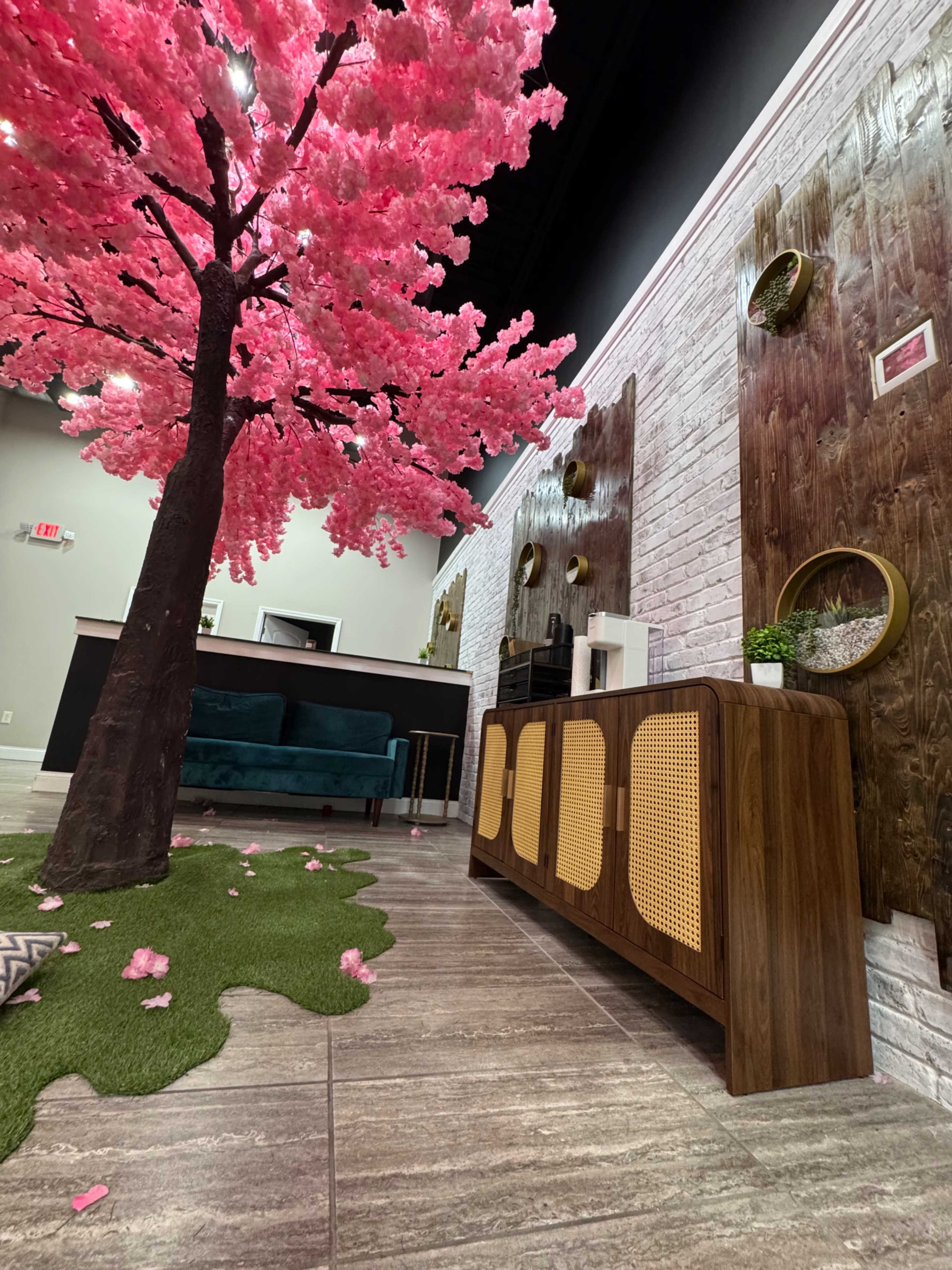 The image features a modern interior space with a large artificial pink cherry blossom tree, a wooden cabinet, and greenery on the floor.