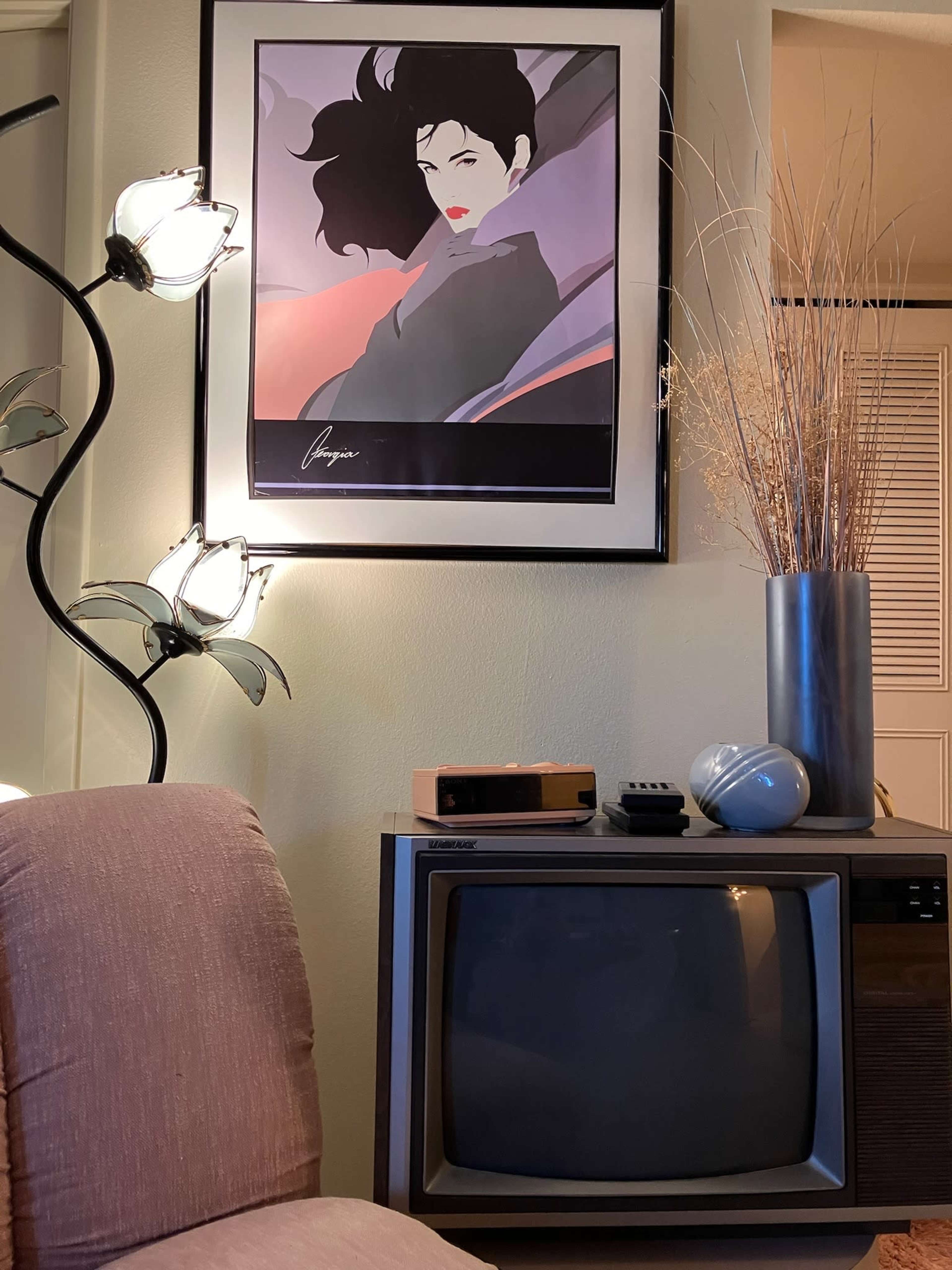 The image shows a vintage television set placed next to a modern lamp with floral motifs, a decorative vase with dried twigs, and an art print of a woman in bold colors hanging on the wall.