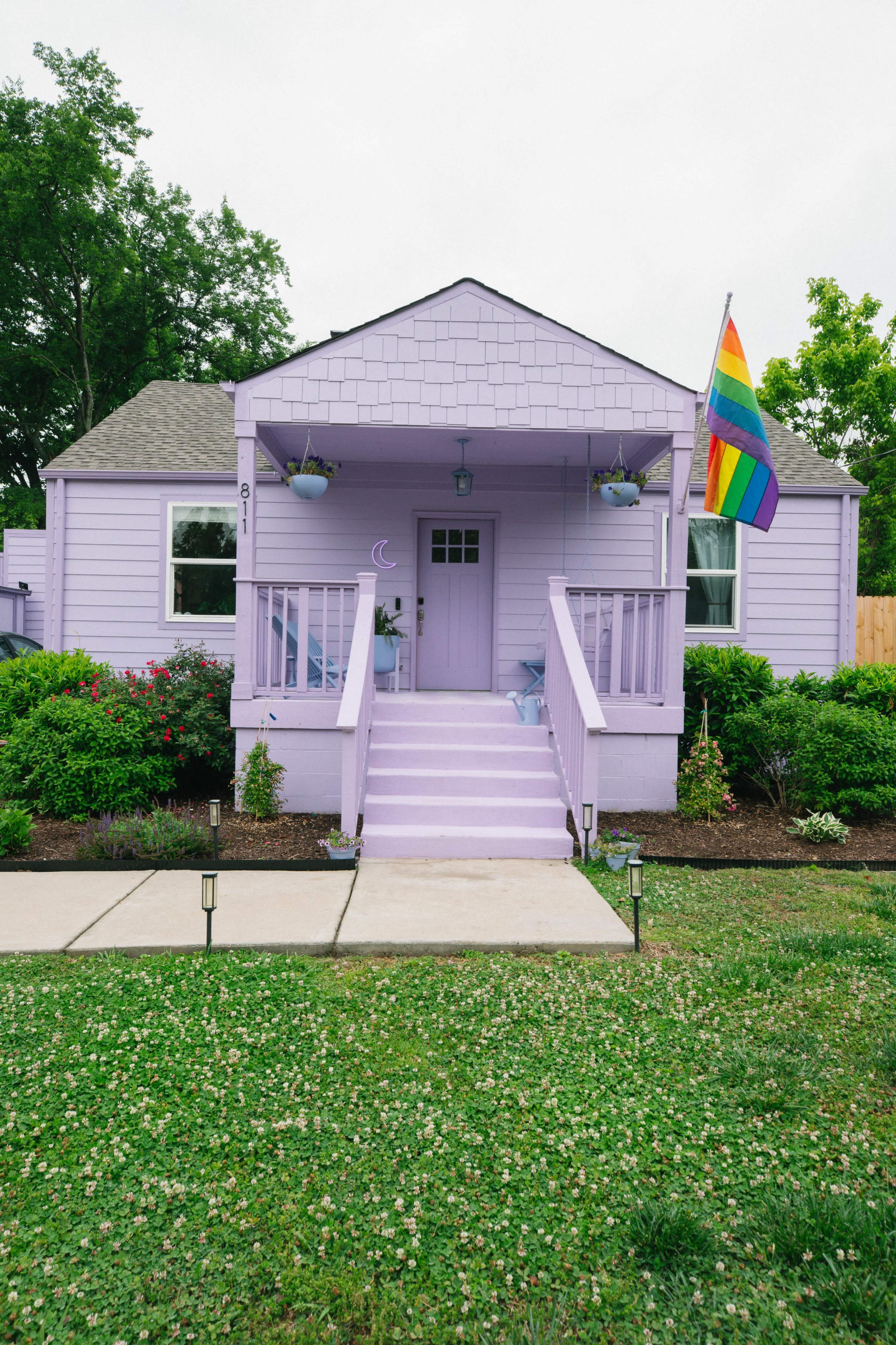 A pastel purple house with a front porch, surrounded by greenery, displays a rainbow flag on a pole.