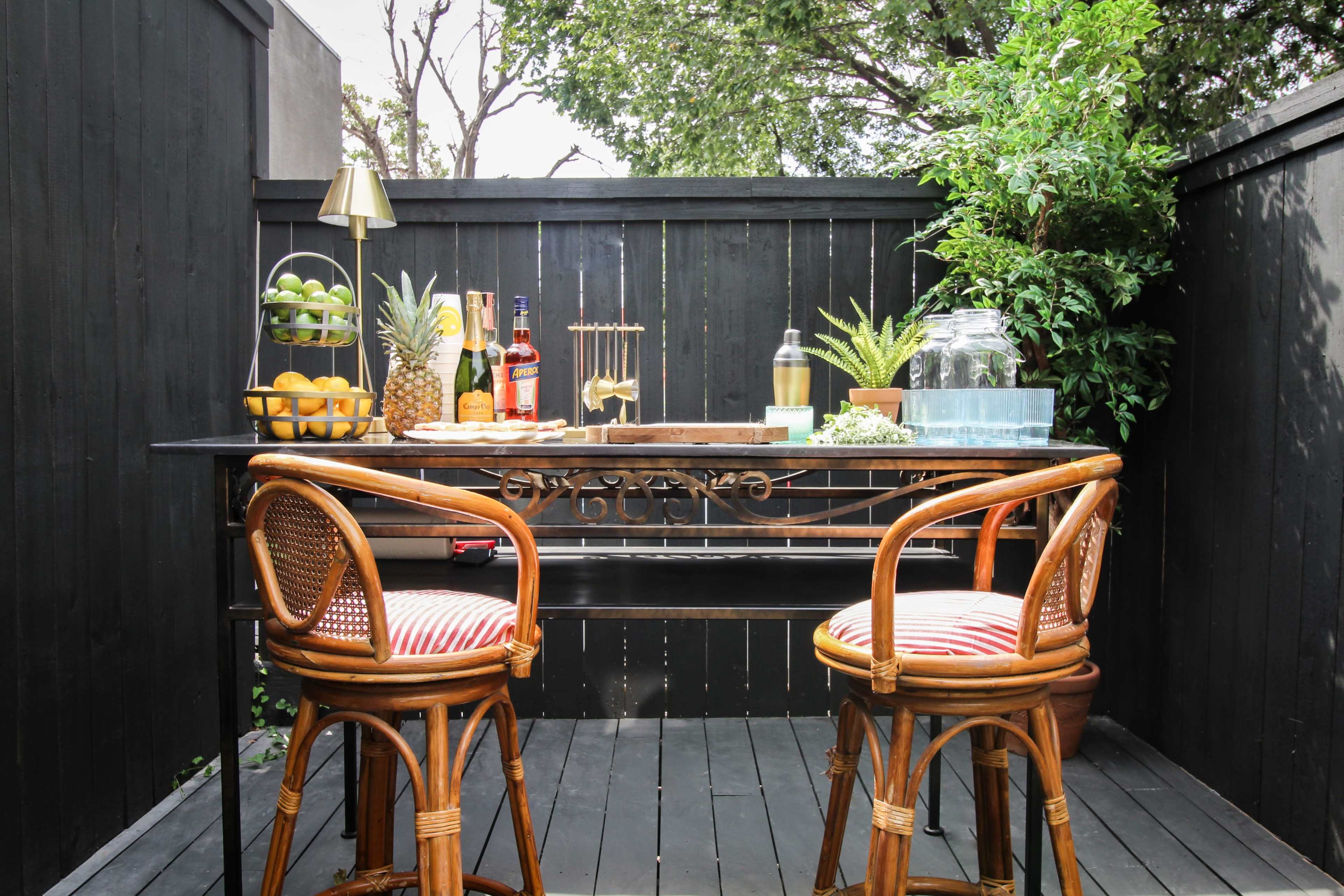 A small outdoor space features a black wooden table with two rattan chairs, surrounded by greenery and containing various drinks and fruit displays.