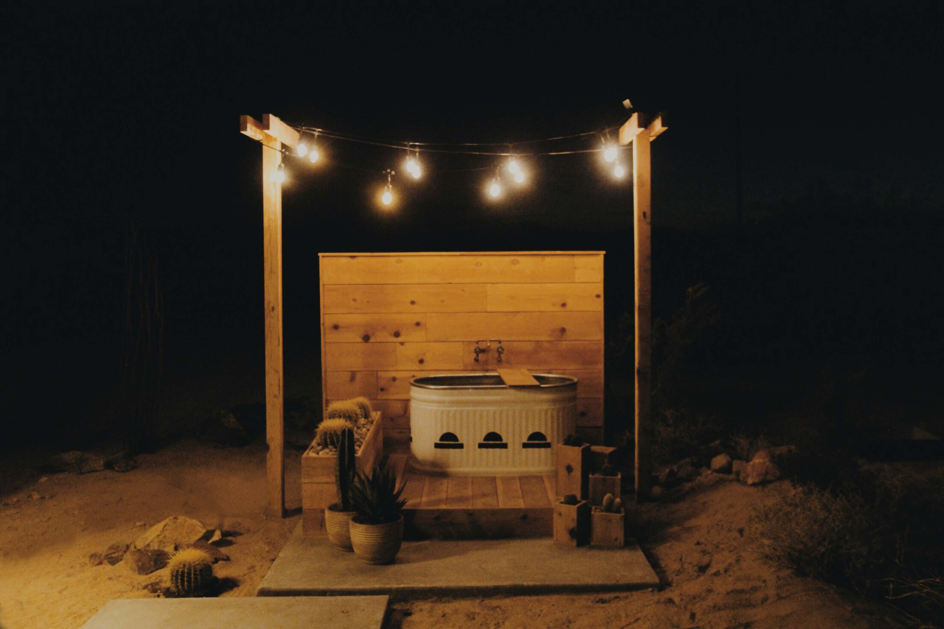 An outdoor bathtub area is illuminated by string lights against a wooden backdrop in a desert setting at night.