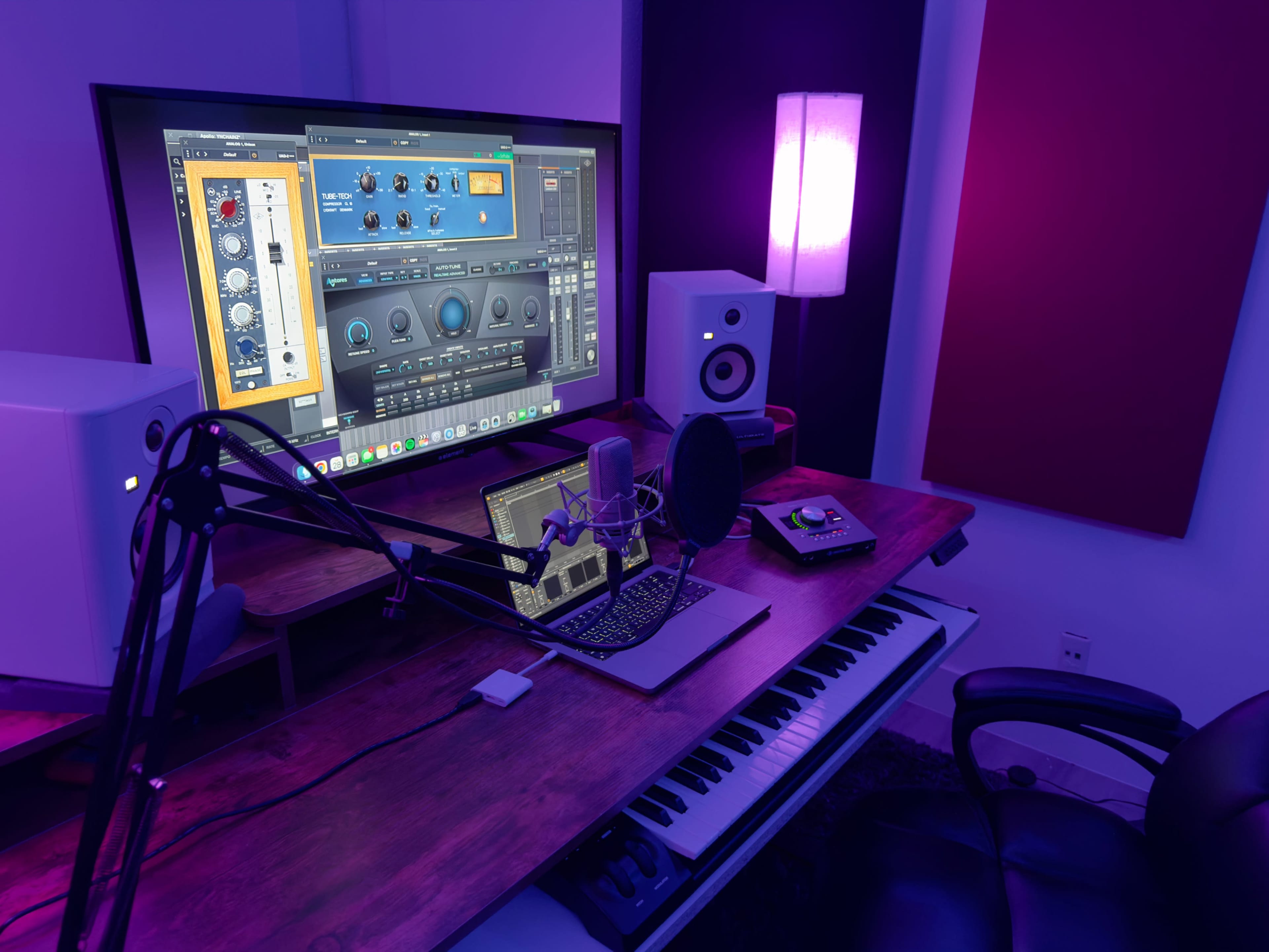 A music production workspace features a computer monitor displaying audio software, a microphone on a stand, speakers, a laptop, and a keyboard under purple and blue lighting.