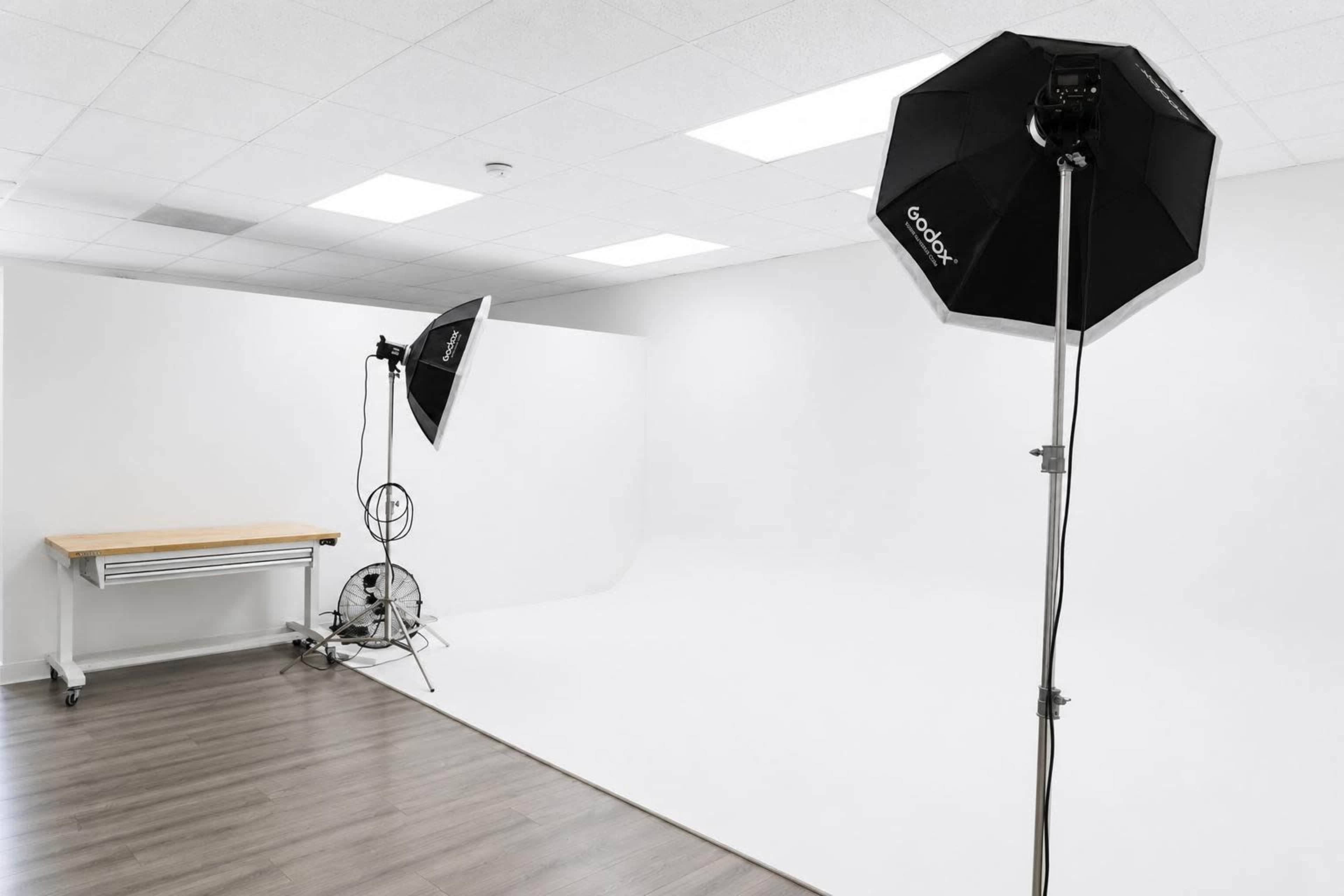 The image shows a photography studio with two softbox lights and a wooden table against a white backdrop.