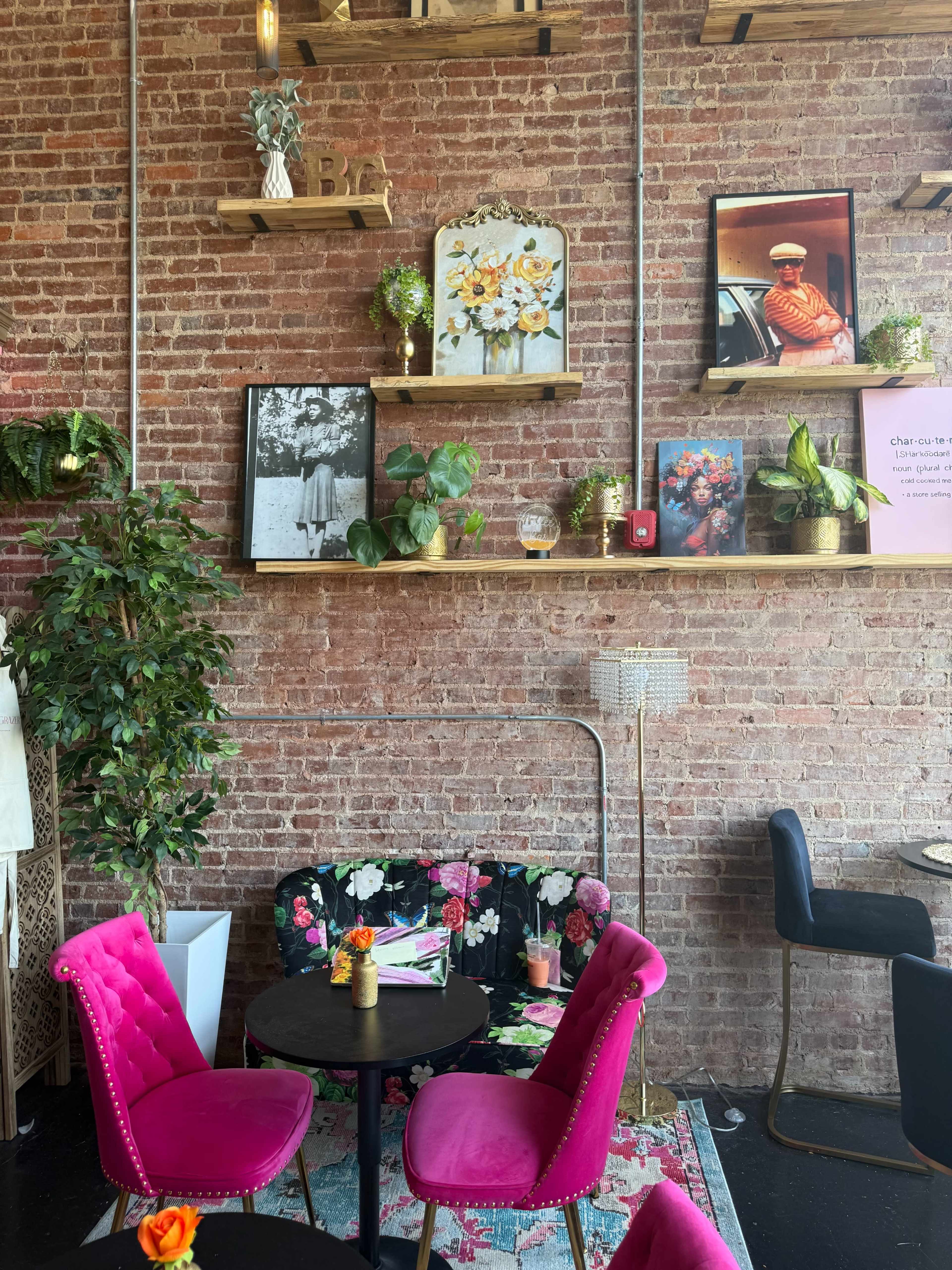 The image shows a cozy café interior with pink chairs, a small round table, and brick walls adorned with plants and various framed artworks.