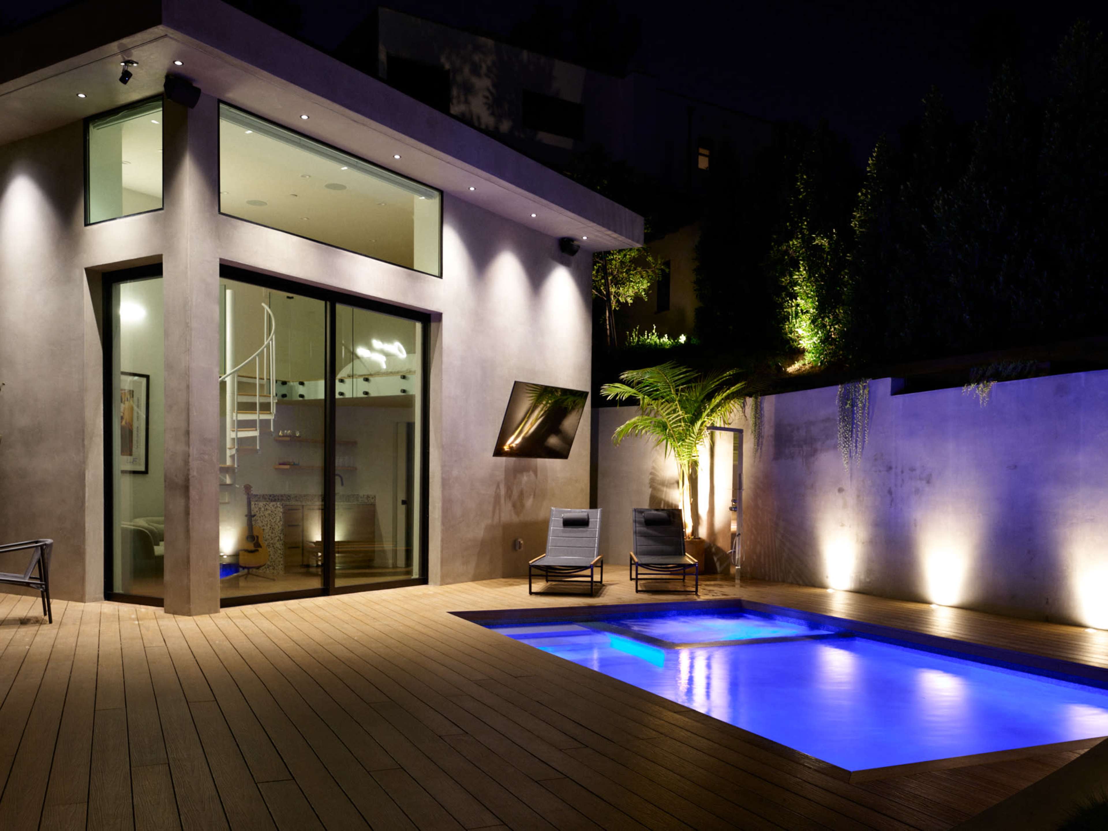 A modern house with large glass windows, a small illuminated pool, and two lounge chairs on a wooden deck at night.