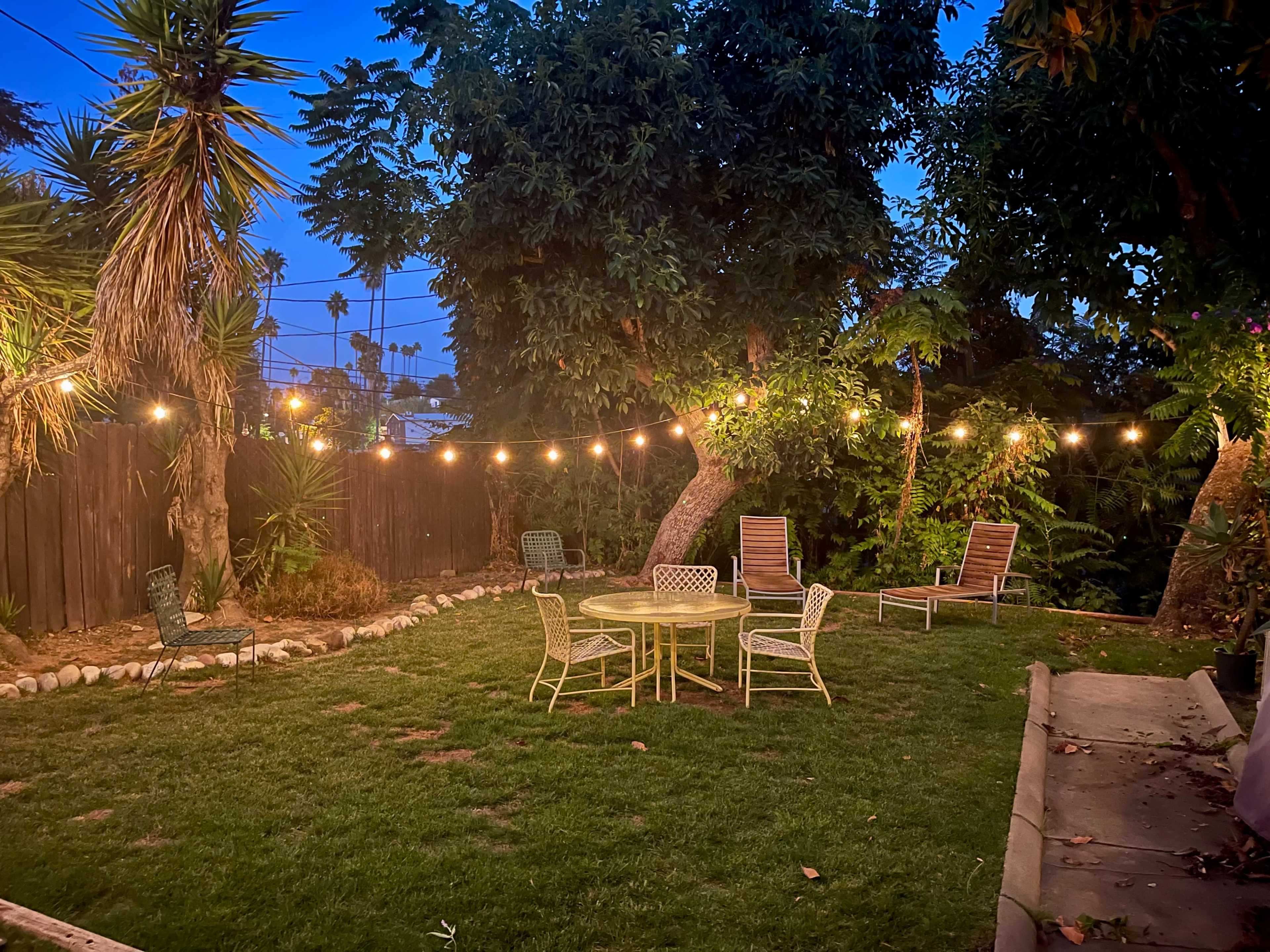 A well-lit backyard features a circular table surrounded by four chairs, with string lights illuminating the scene and palm trees in the background.