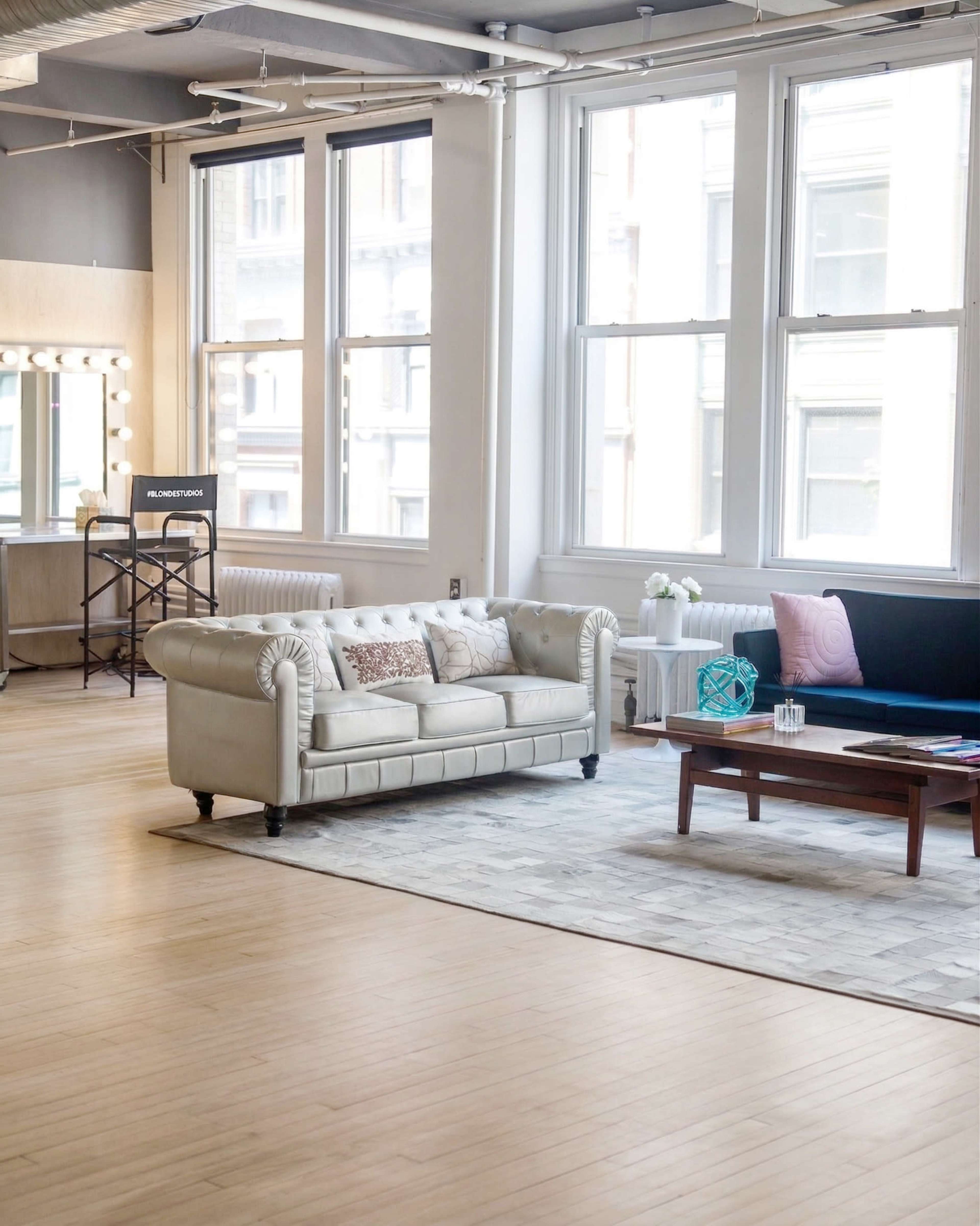 The image shows a spacious living area with a light-colored couch, a dark sofa, a wooden coffee table, and large windows allowing sunlight to enter.