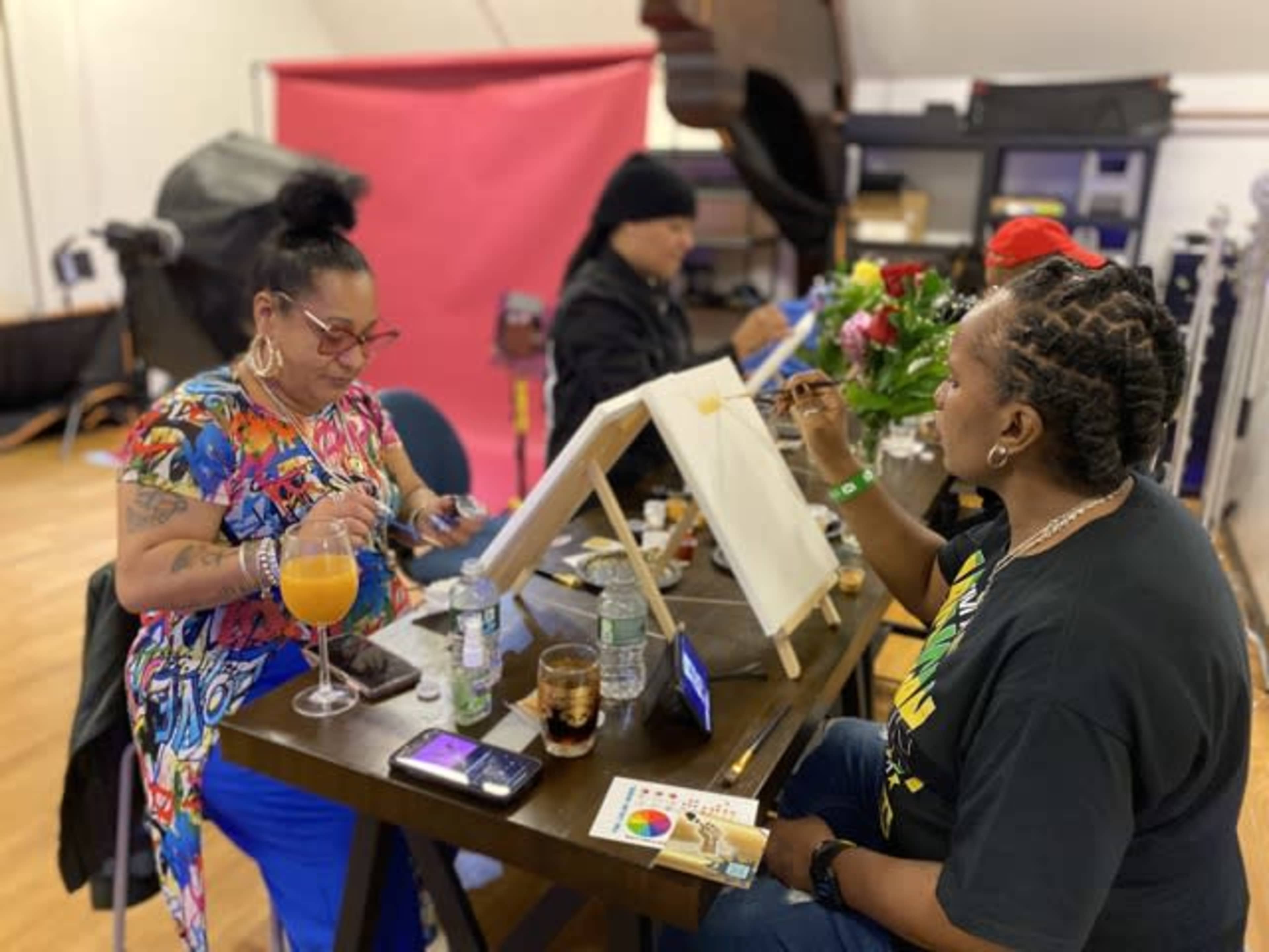 A group of four people are engaged in a painting activity in a studio, with two individuals painting on easels and others socializing nearby.
