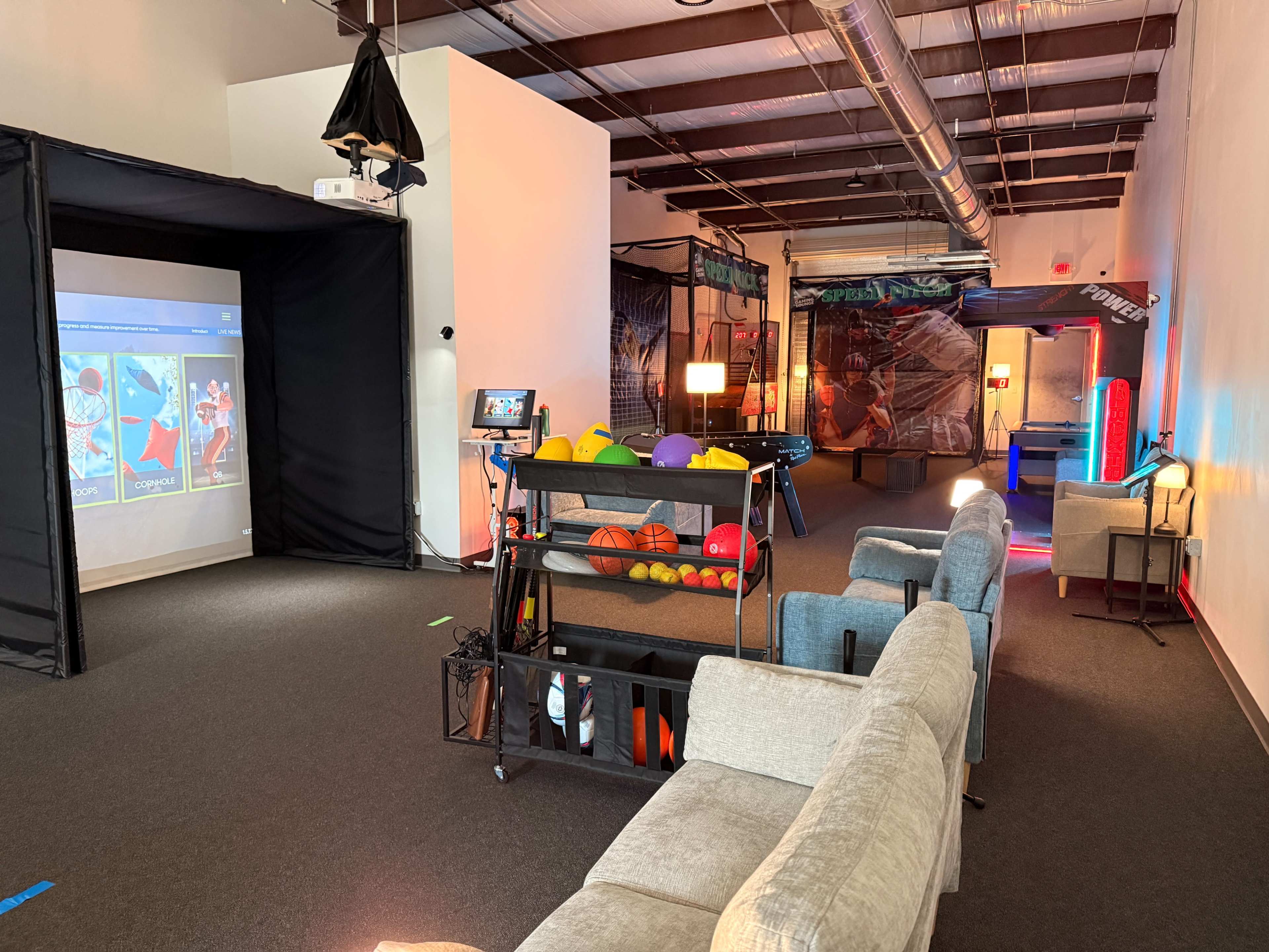The image shows a recreation space with gaming stations, comfortable seating, and various sports equipment arranged in an open area.