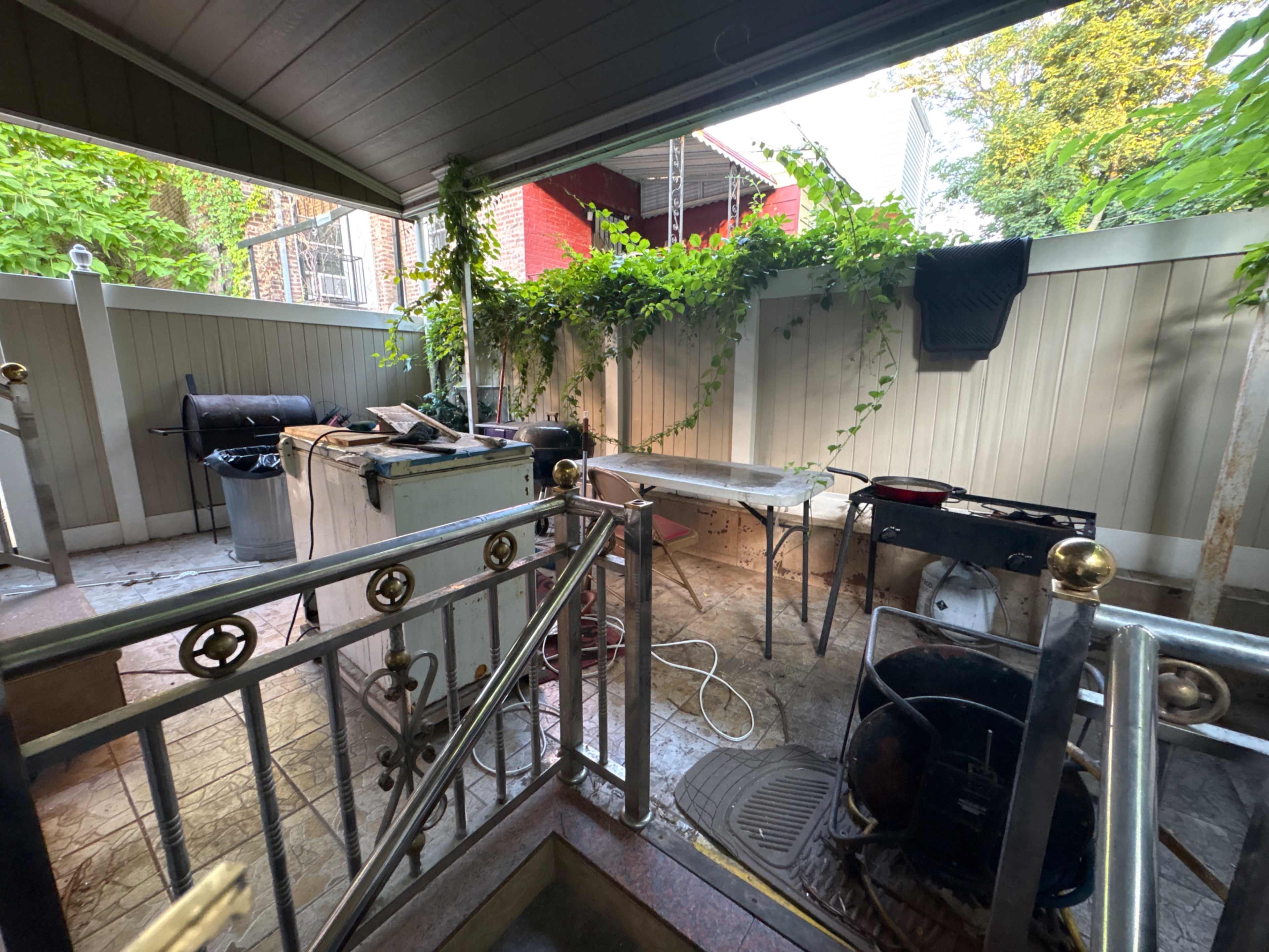 The image shows a cluttered outdoor space with a mixture of cooking equipment, tools, and greenery along the walls.