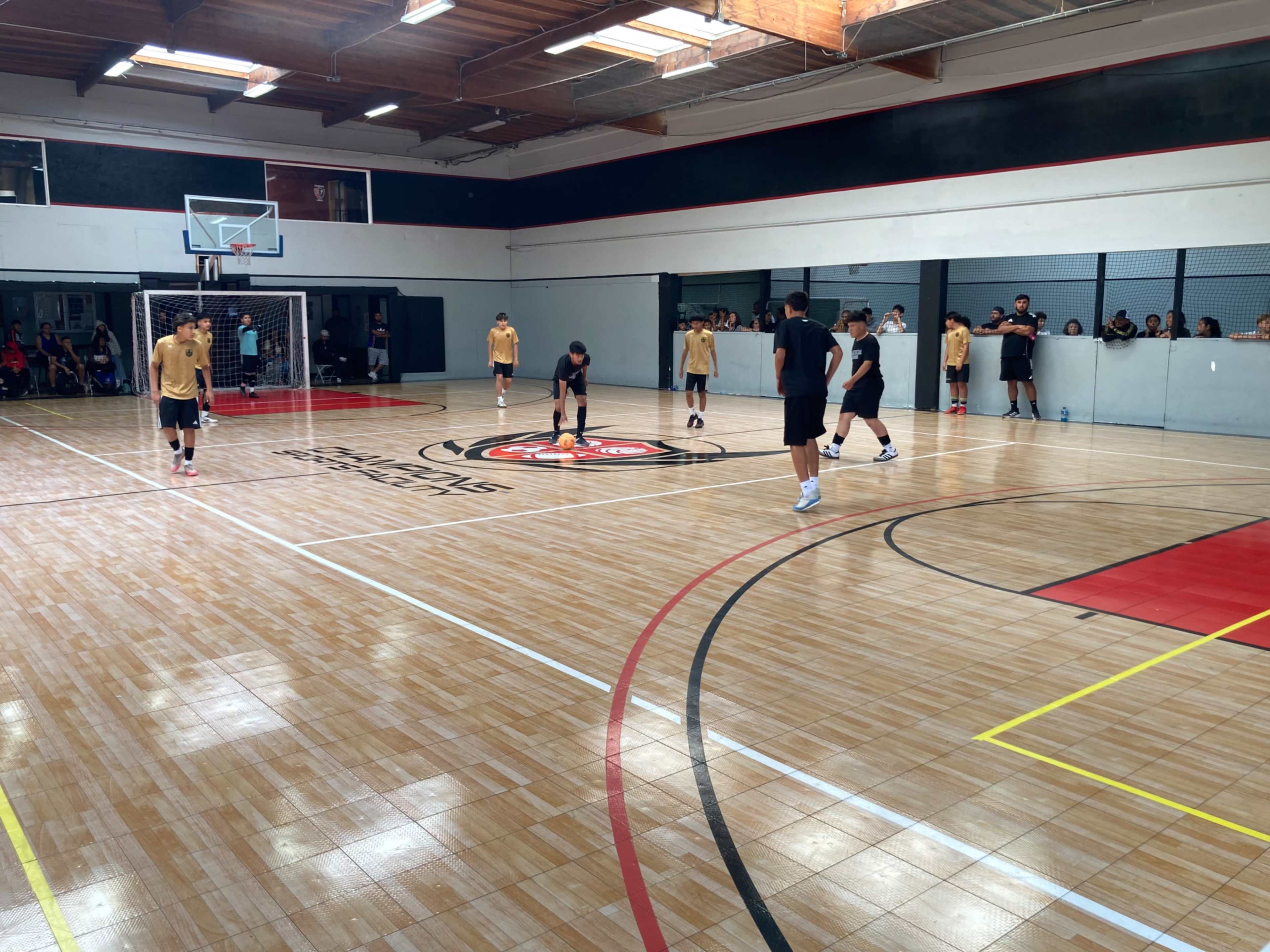 A group of players in yellow and black uniforms practices on a wooden gym floor marked for sports, with spectators observing from the sidelines.