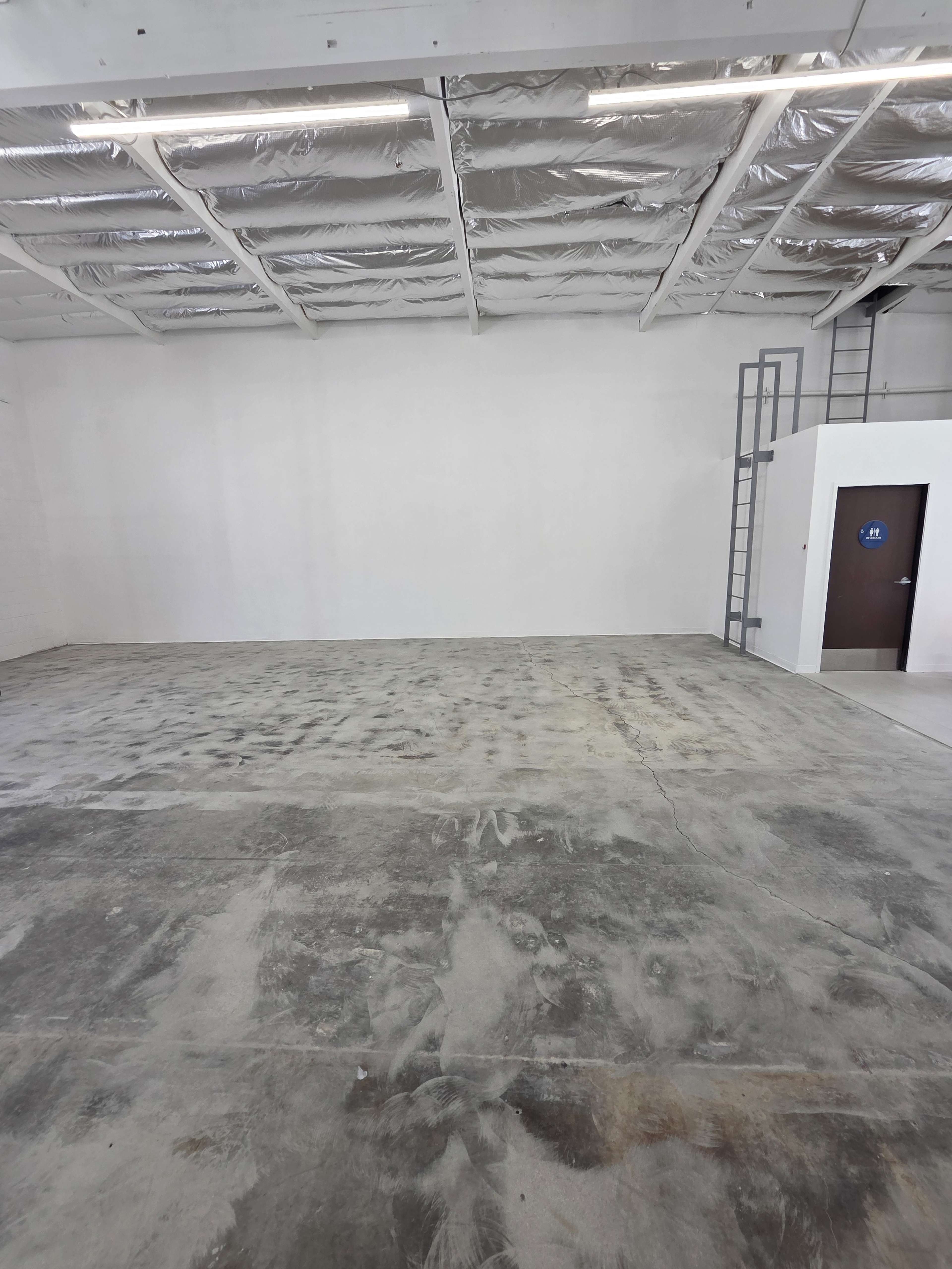 The image shows a spacious, empty industrial room with concrete flooring and white walls, featuring a single door and a ladder against one wall.