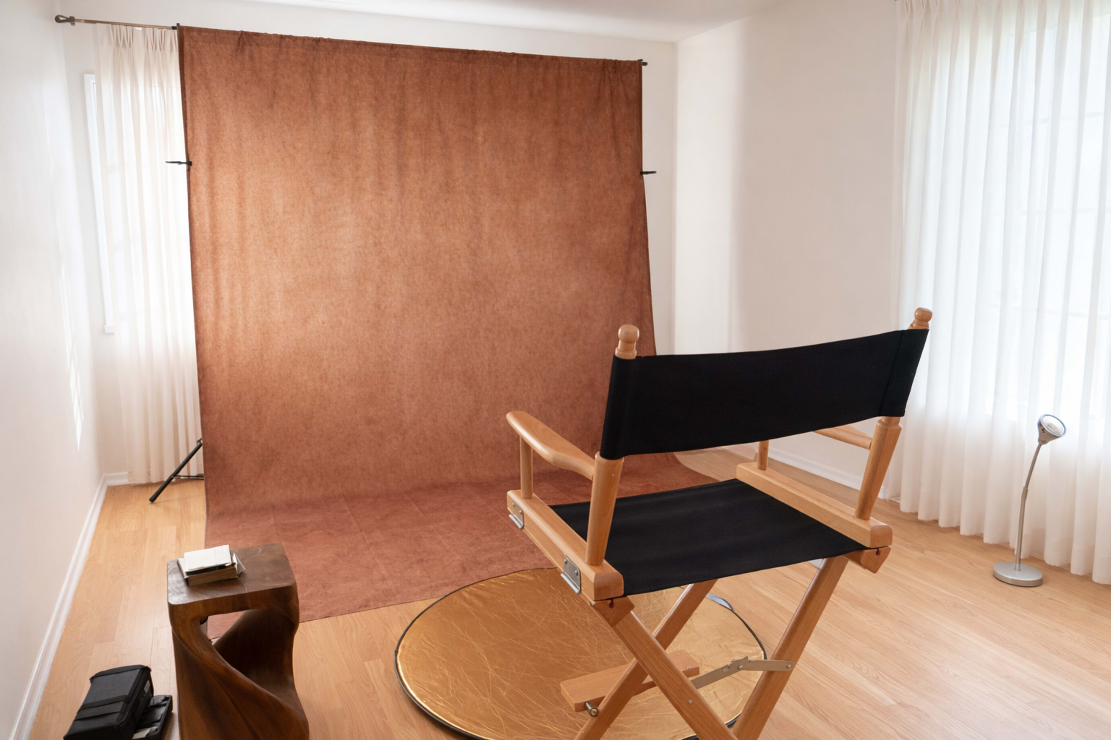 The image shows a simple studio setup featuring a director's chair facing a brown backdrop with soft natural light filtering through white curtains.