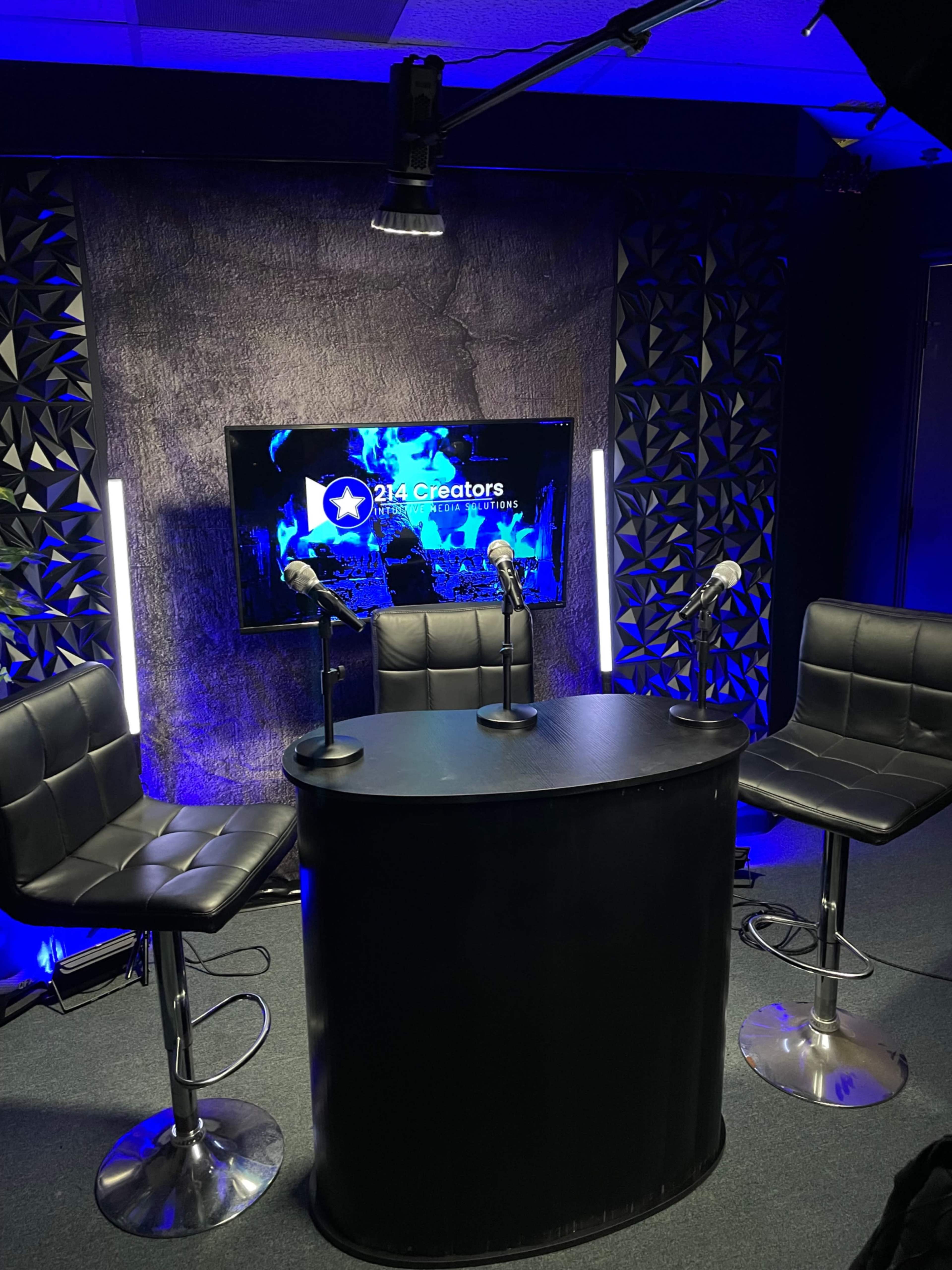 A modern studio setup with three barstools and a circular table in front of a backdrop featuring a screen that displays "214 Creators."