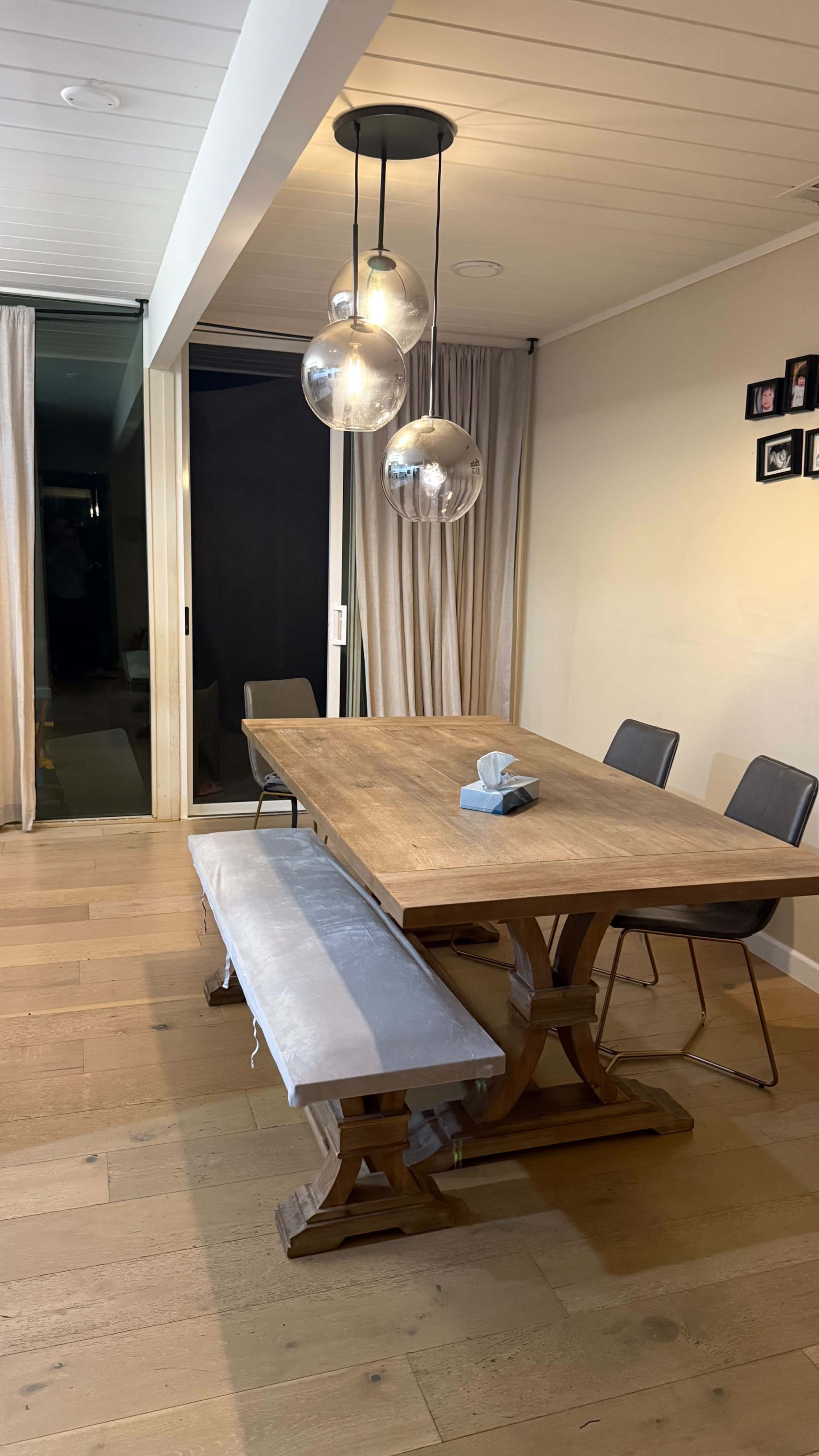 The image shows a dining area featuring a wooden table with a bench and chairs, illuminated by three round pendant lights.