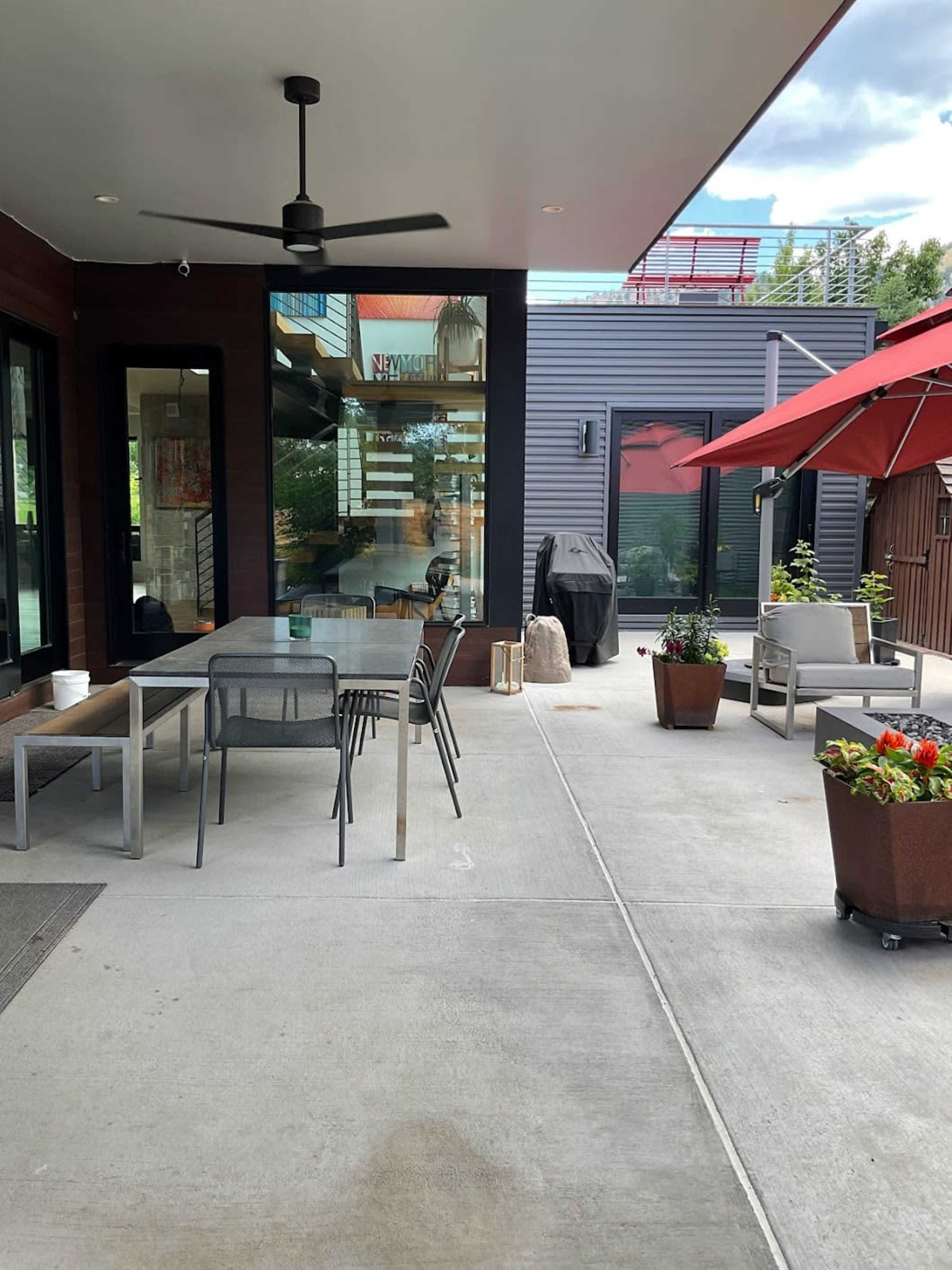 The image shows a modern outdoor patio area featuring a table with chairs, a red umbrella, potted plants, and a grill.