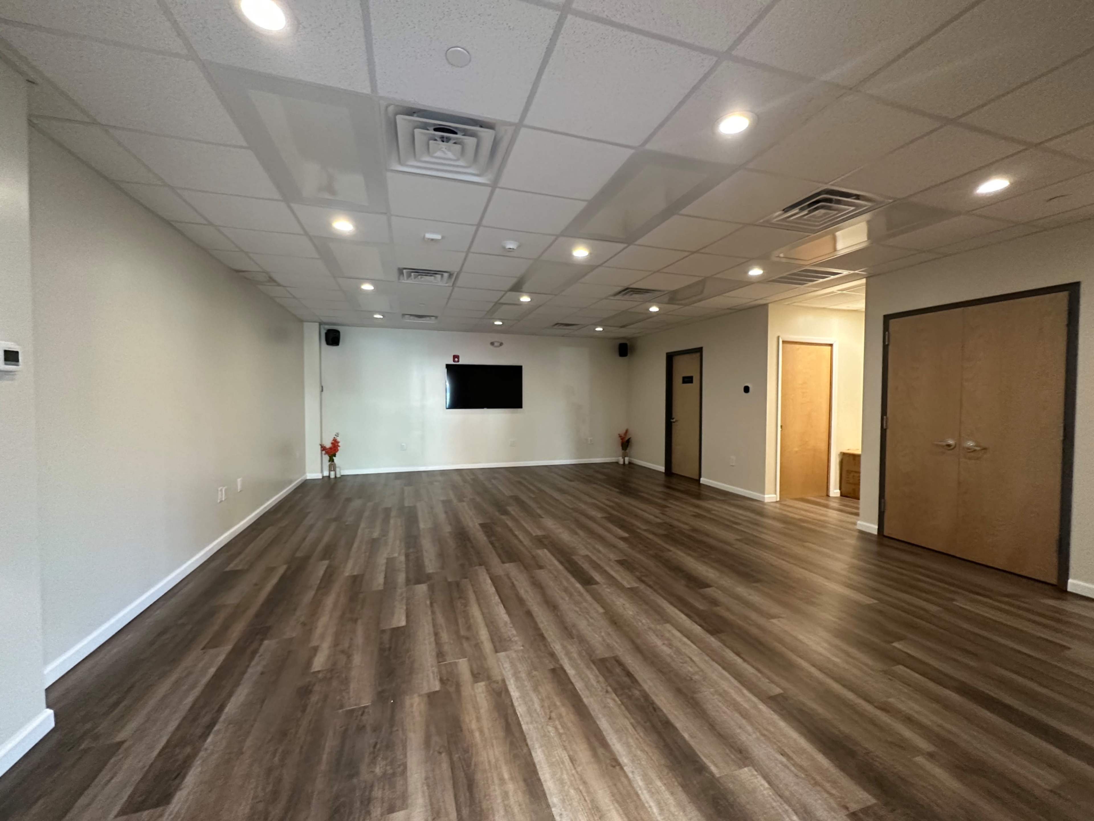 A spacious, empty room with hardwood flooring, a flat-screen TV mounted on the wall, and two closed doors.