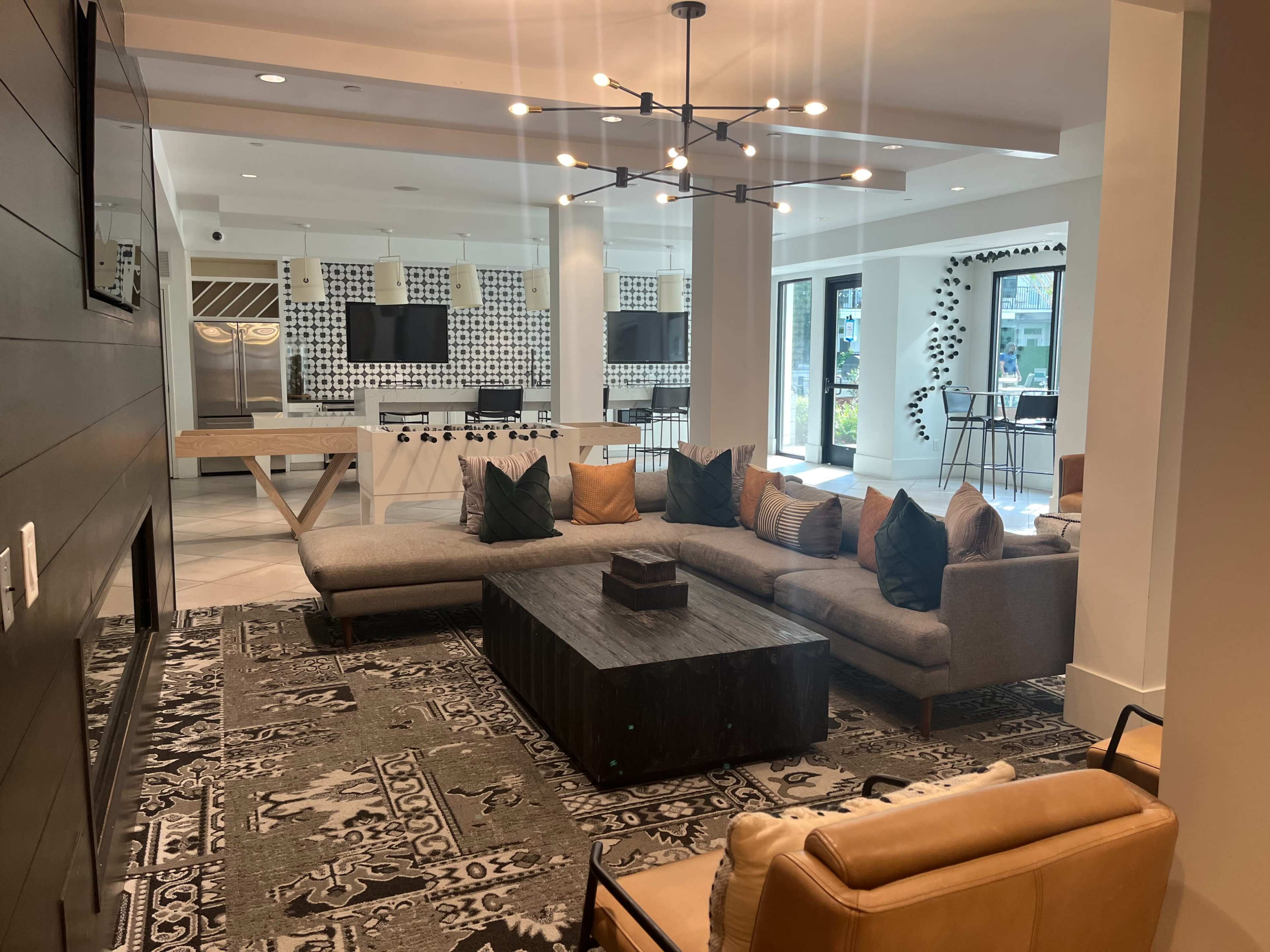A modern lounge area with a sectional sofa, a dark coffee table, and a kitchen in the background, featuring two televisions and decorative wall patterns.