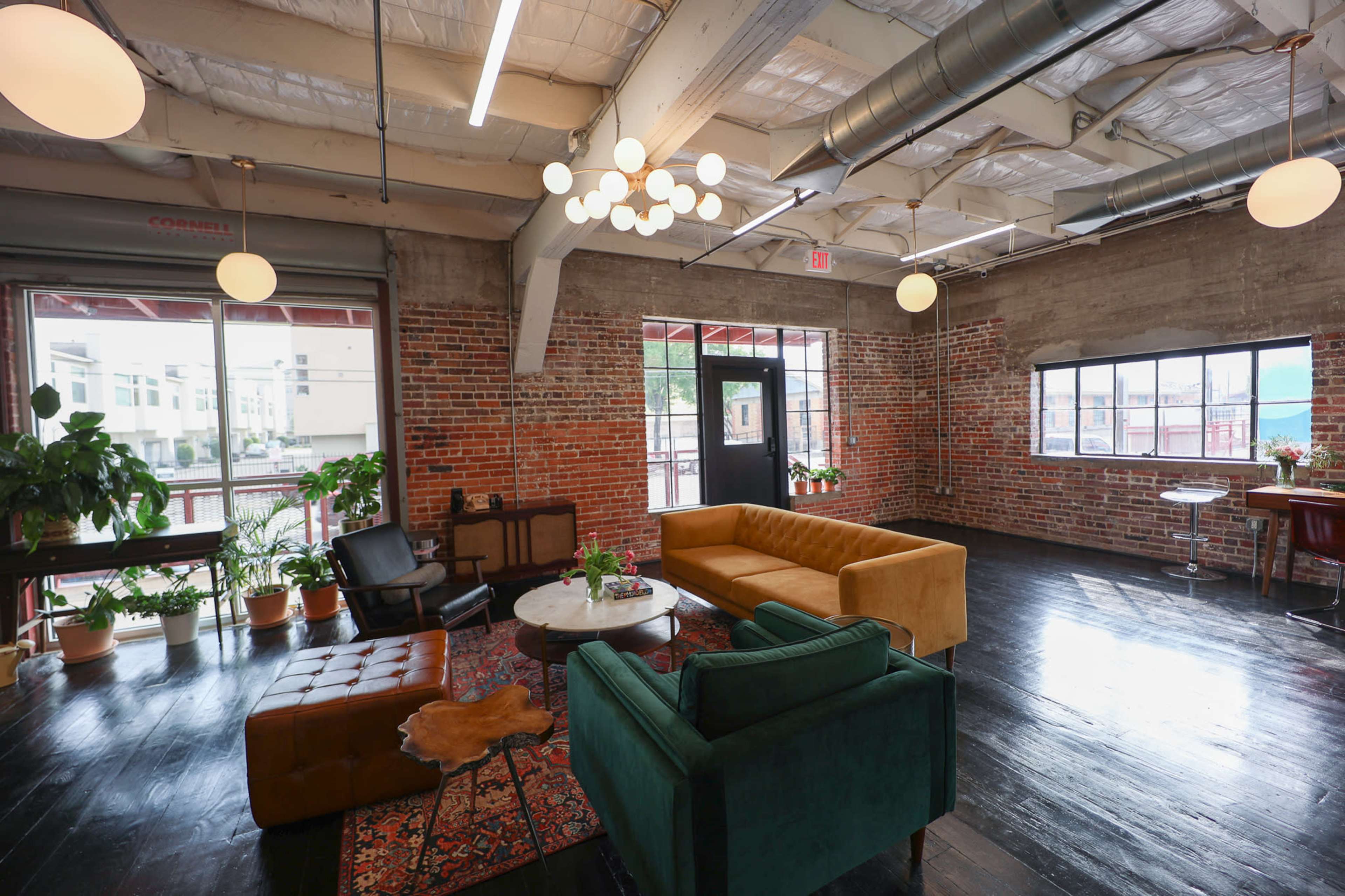 The image shows a spacious, loft-style room with exposed brick walls, modern light fixtures, and a mix of seating arrangements including a mustard-colored sofa, a green armchair, and a small coffee table surrounded by plants.