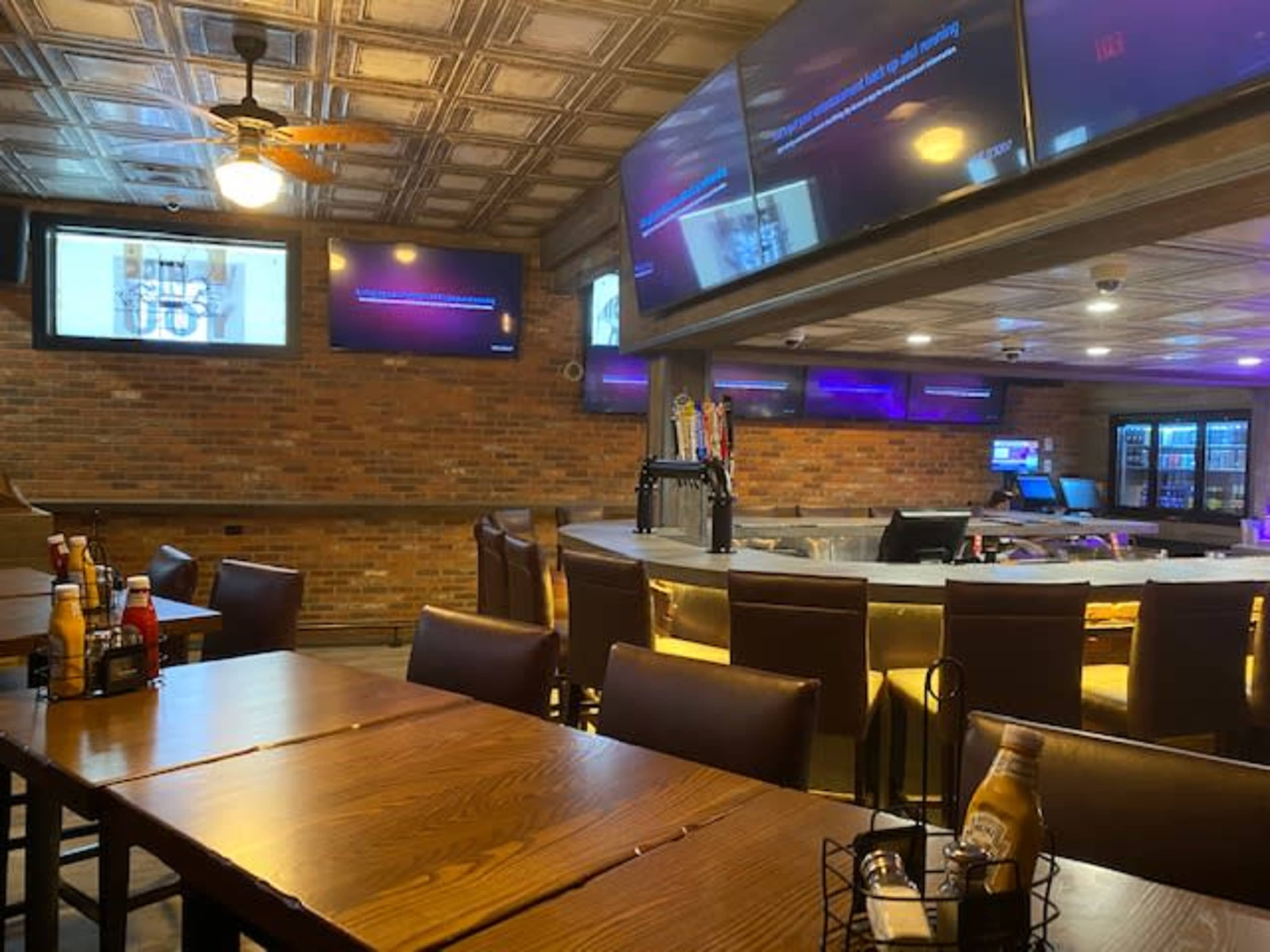 The image shows the interior of a bar or restaurant with wooden tables, leather chairs, a circular bar counter, and multiple screens displaying content on the walls.