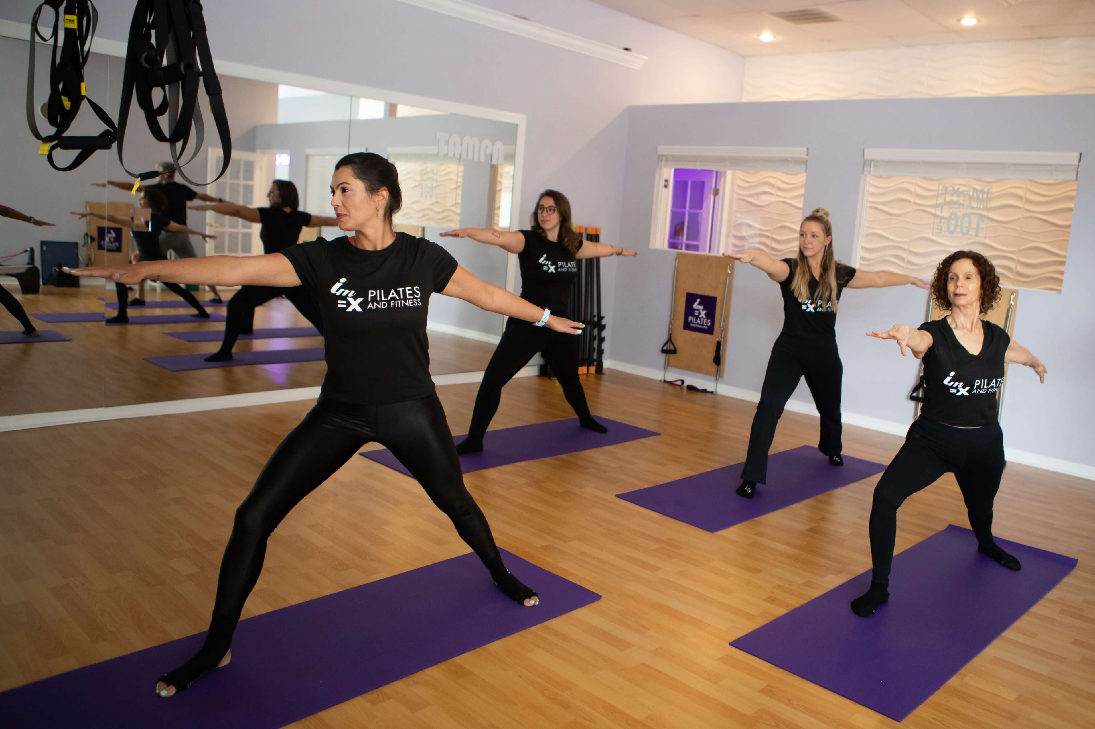 A group of four women performs Pilates exercises on purple mats in a fitness studio.