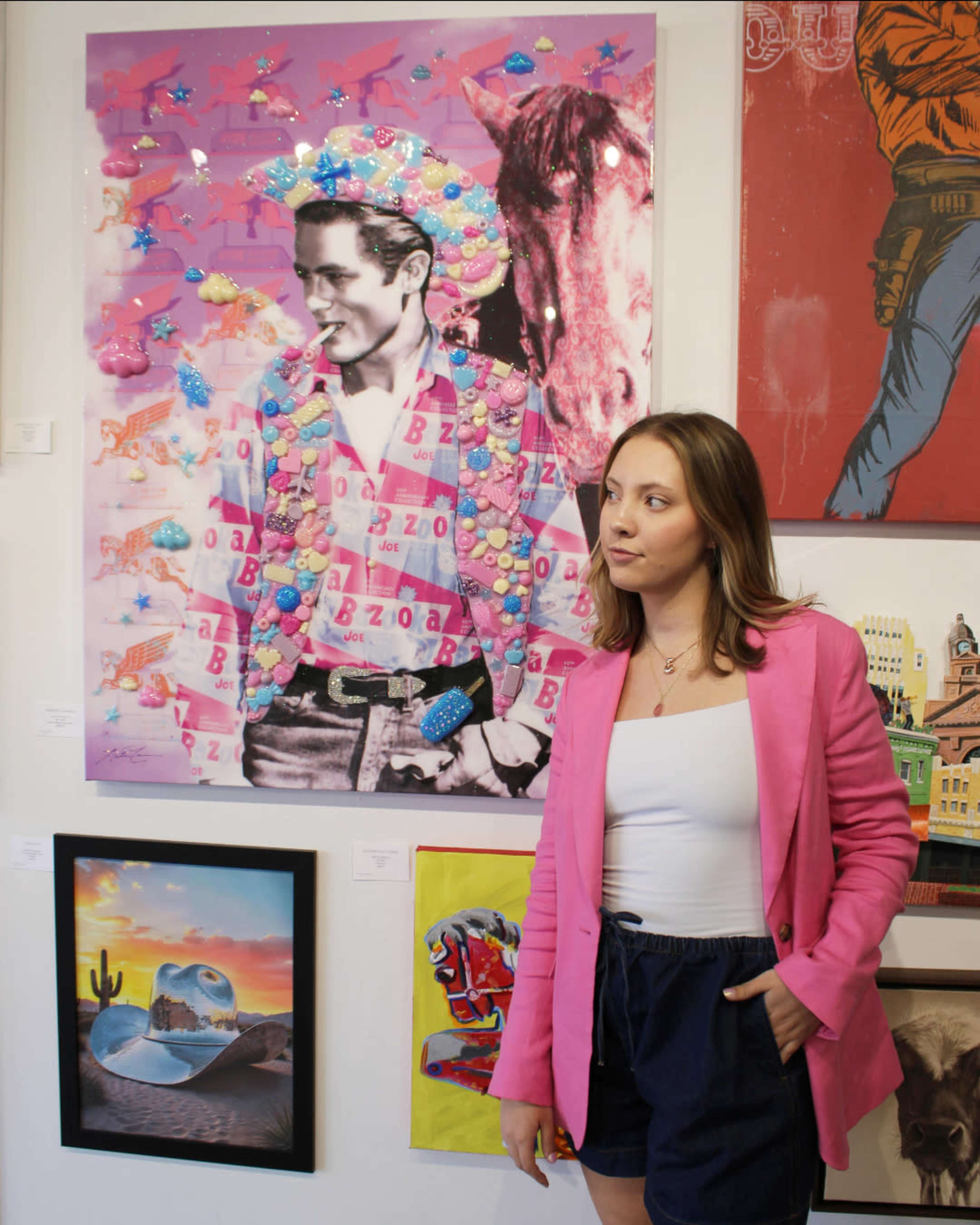 A woman in a pink blazer stands next to a colorful, artistic portrait of a man in a decorative outfit.