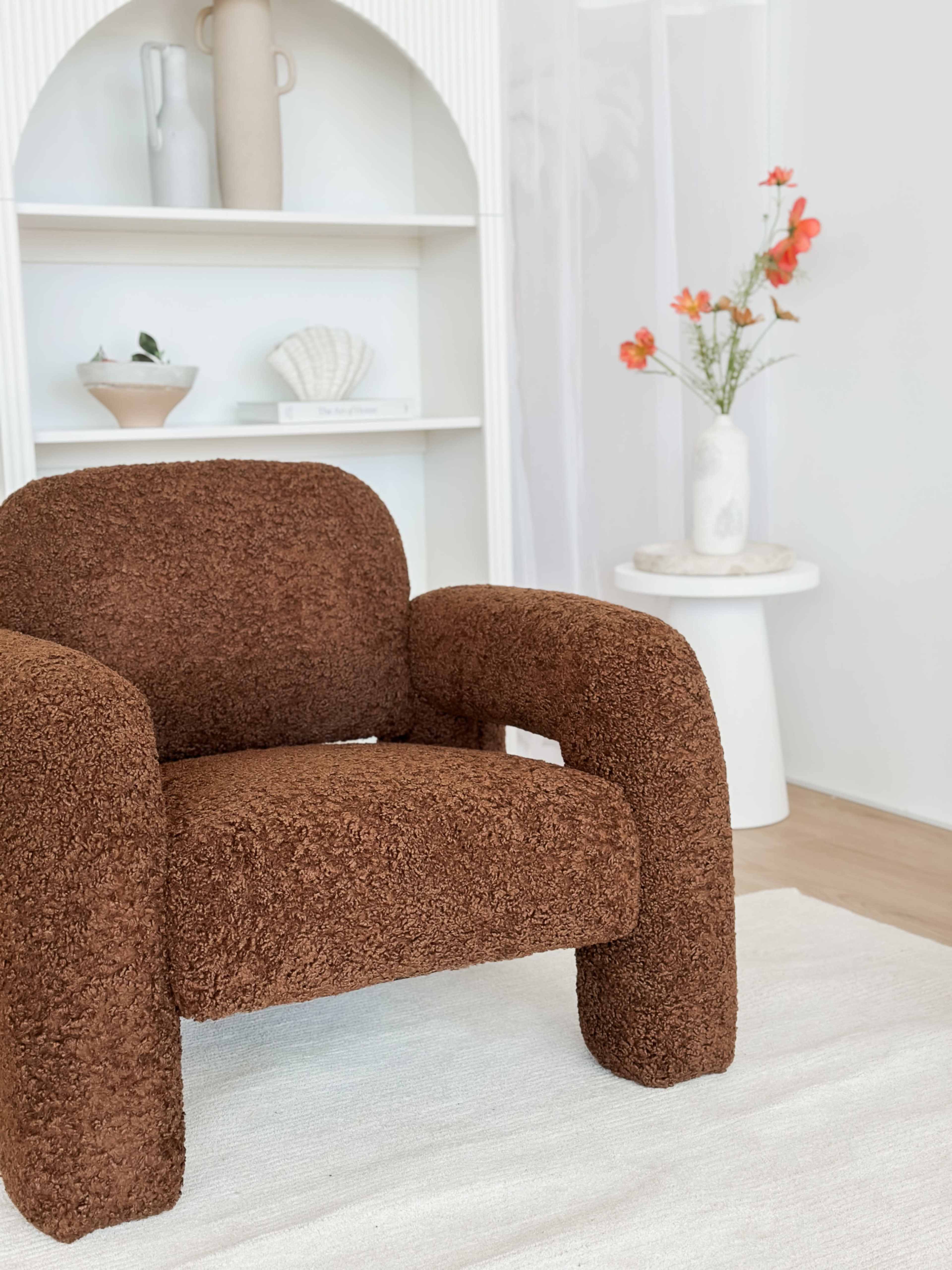 The image features a brown, textured armchair positioned near a white bookshelf and a side table with a vase of flowers.