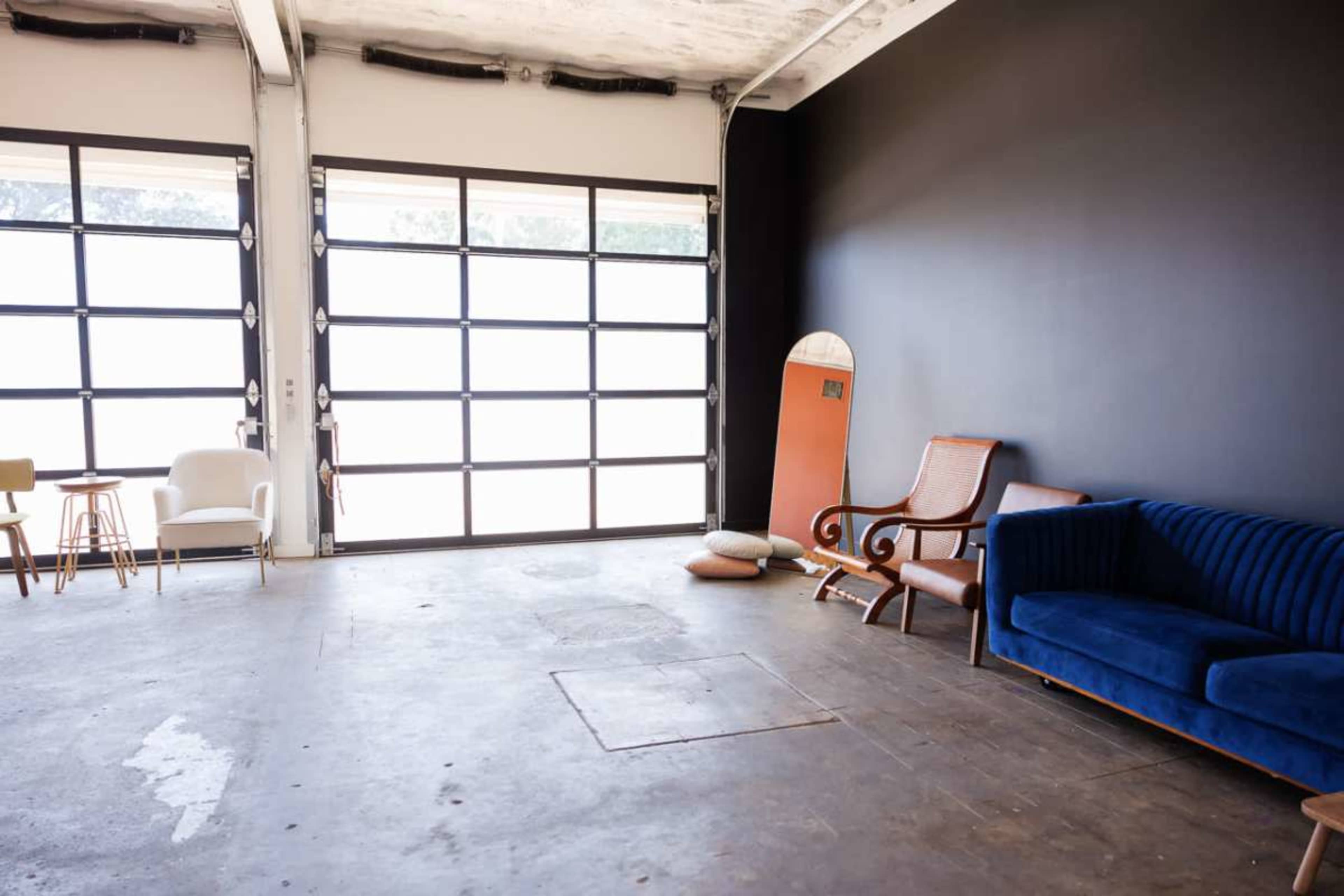 The image shows a spacious room with a concrete floor, featuring a large glass garage door, a blue couch, a wooden chair, a small table, and a surfboard leaning against the wall.