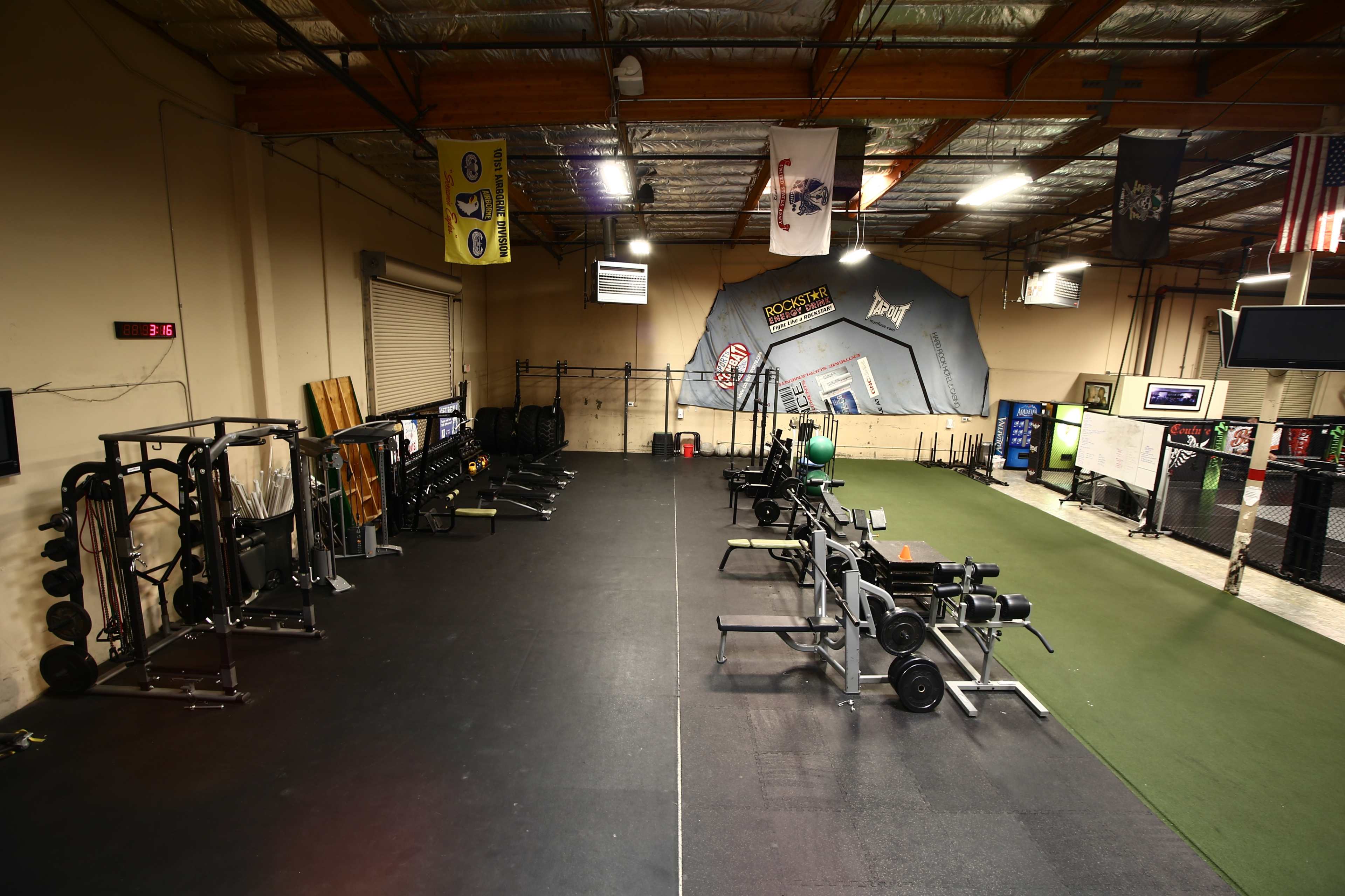 A spacious gym with various weightlifting equipment and exercise areas, separated by a black rubber floor and green turf.