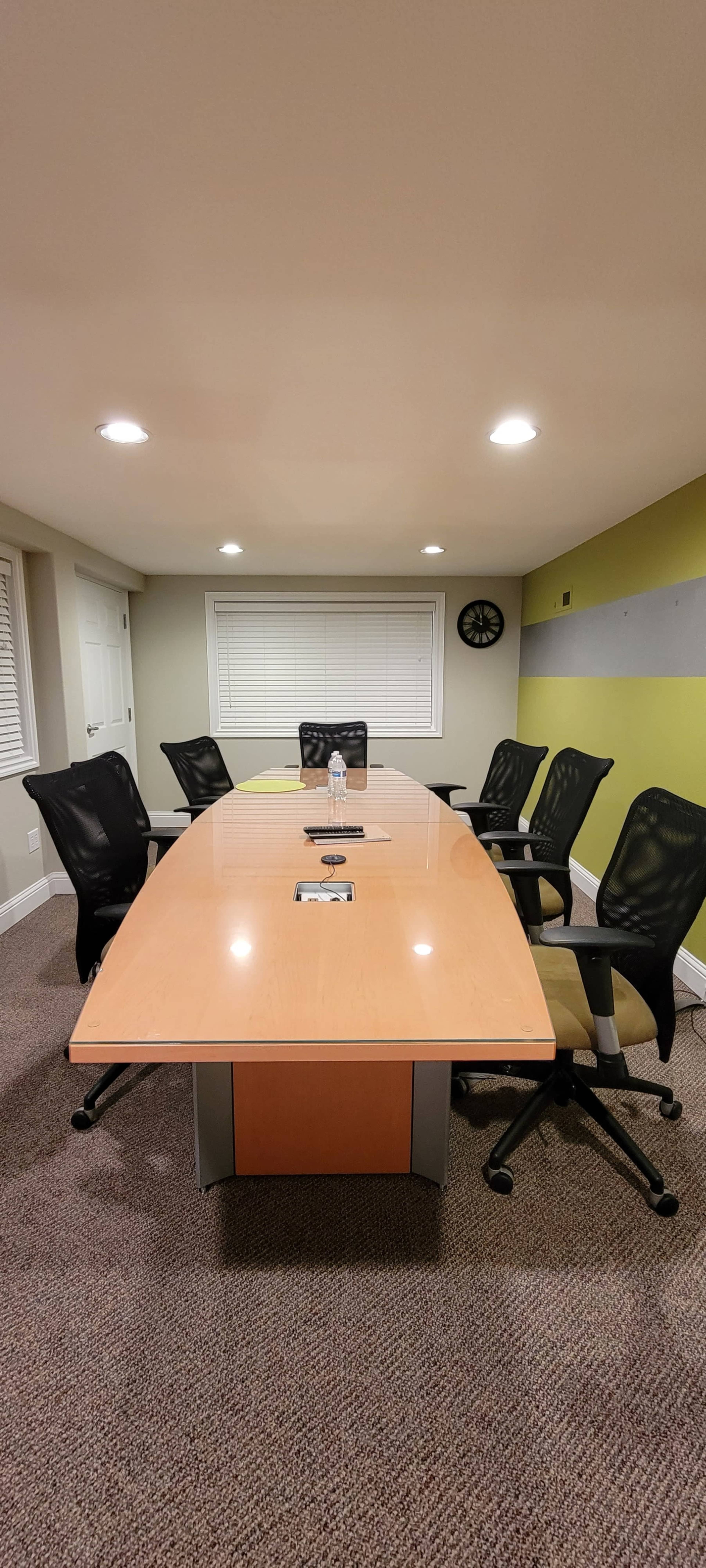 A conference room features a long wooden table with six ergonomic chairs, wall-mounted clock, and a water bottle in the center.
