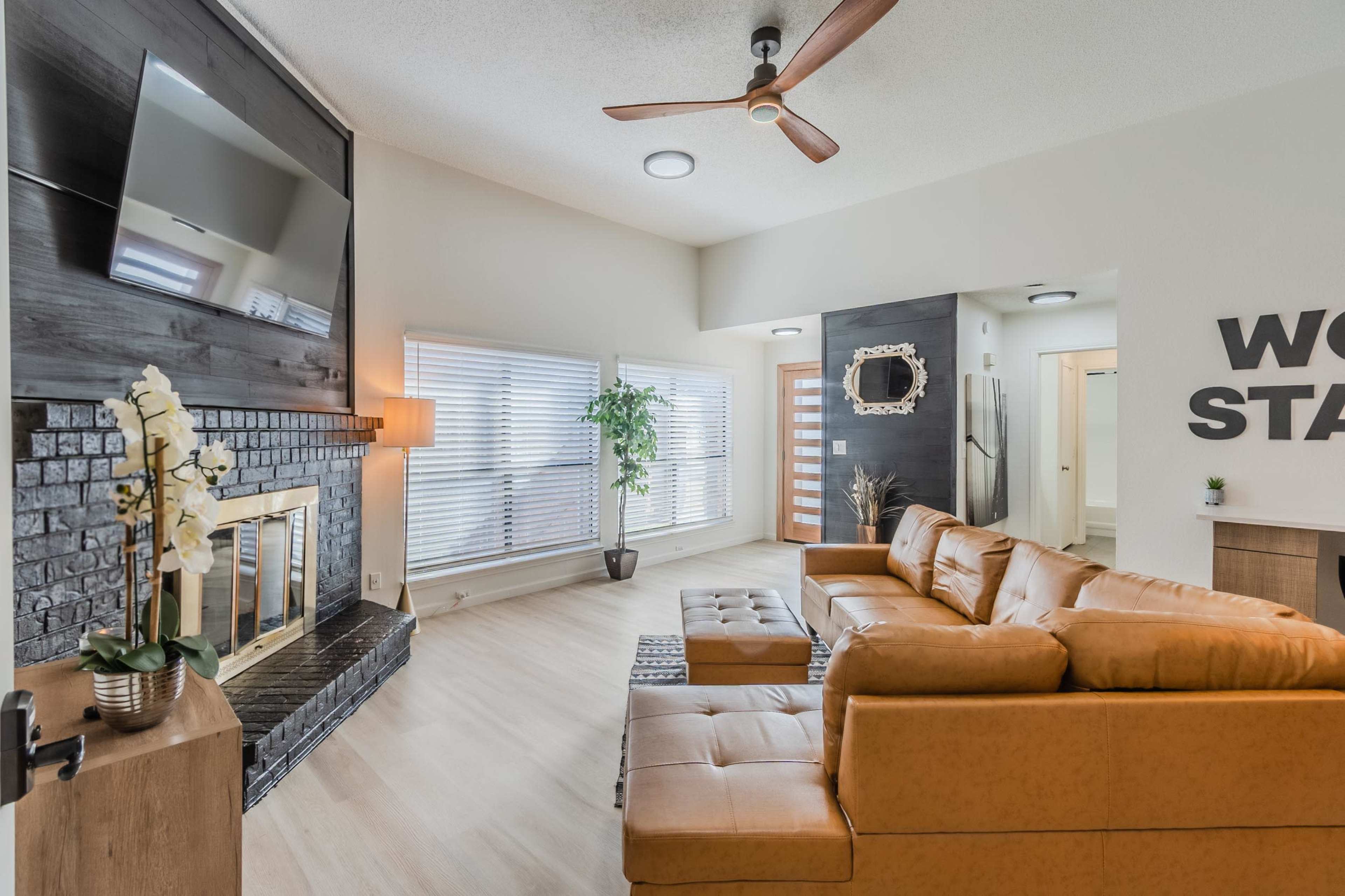 A modern living room features a tan leather sectional sofa, a fireplace with a black surround, and large windows with blinds.
