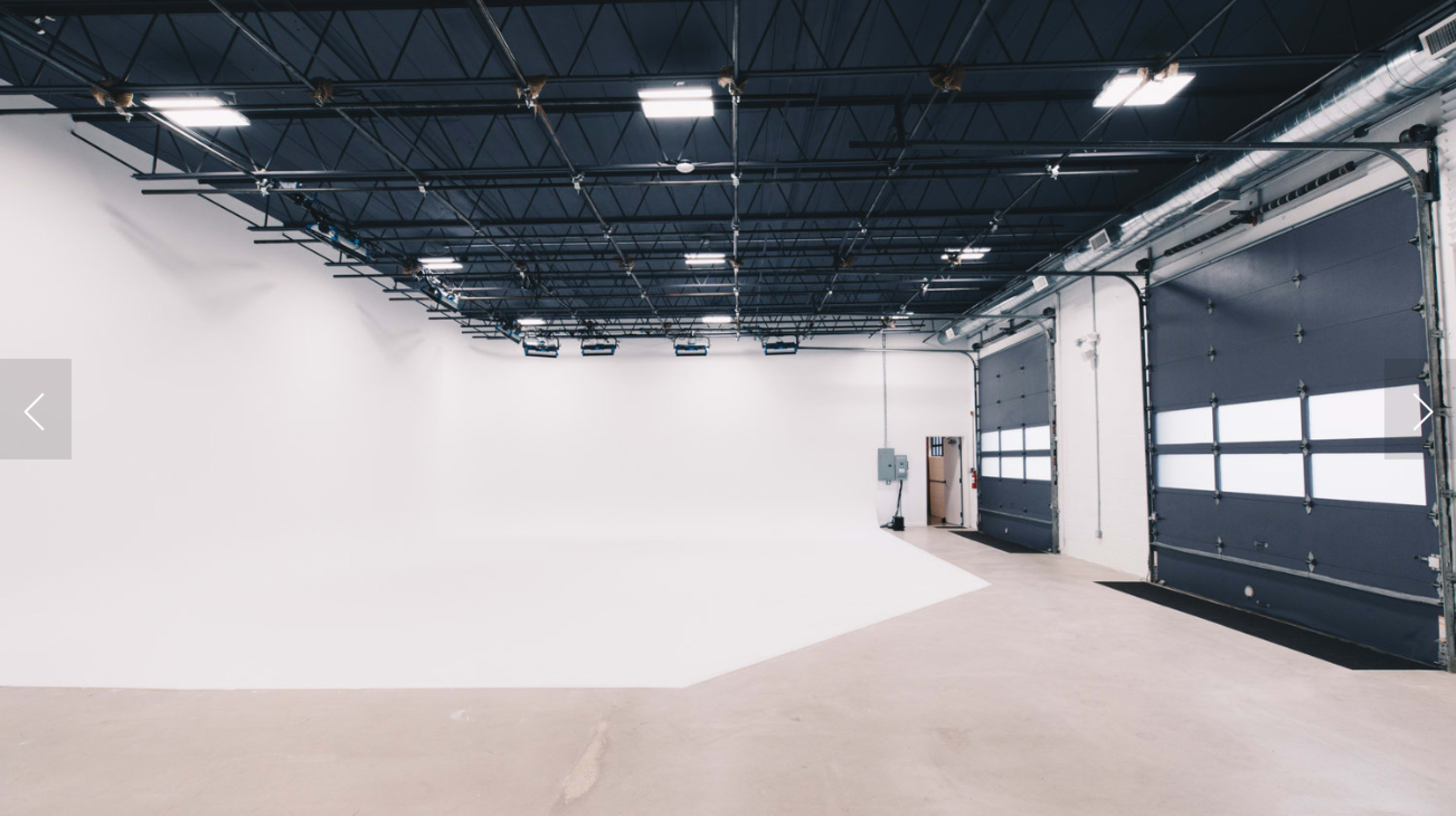 A large, empty studio space features a white backdrop and industrial lighting, with a pair of overhead doors on one side.