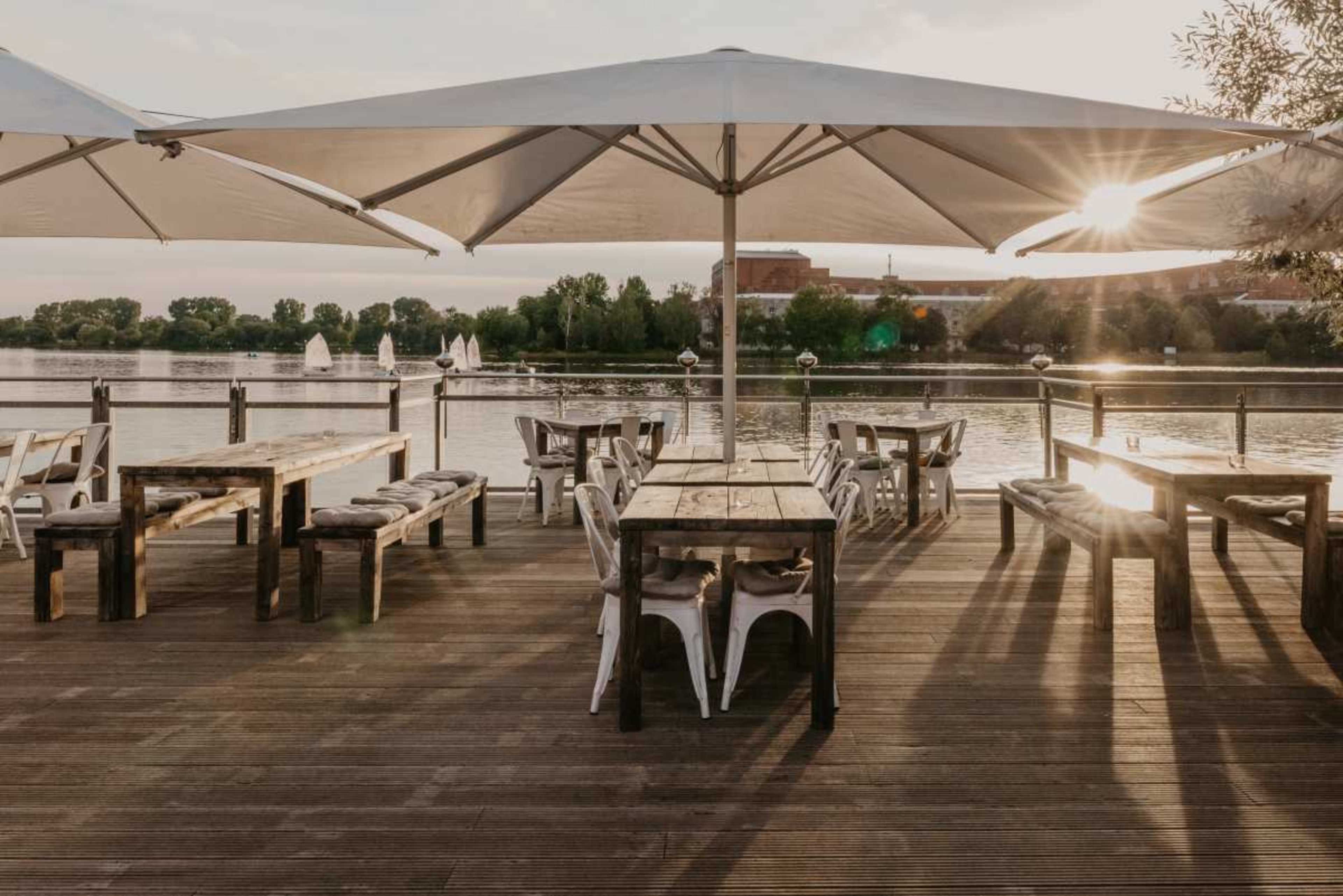A wooden deck with tables and chairs overlooks a river, bordered by large umbrellas and green trees.