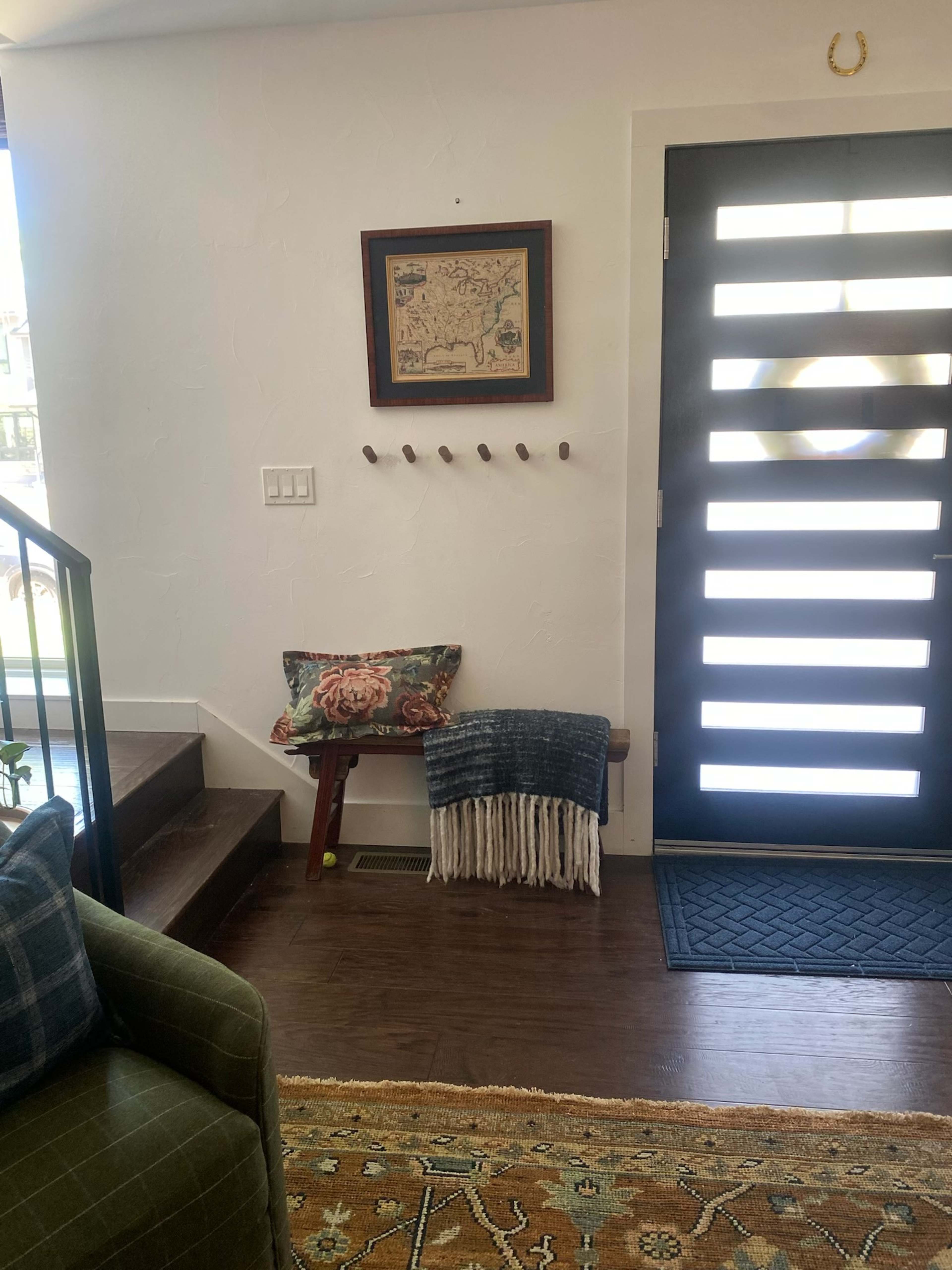 The image shows an entryway featuring stairs, a decorative wall hanging, coat hooks, a small bench with a throw blanket, and a modern front door with horizontal slats.
