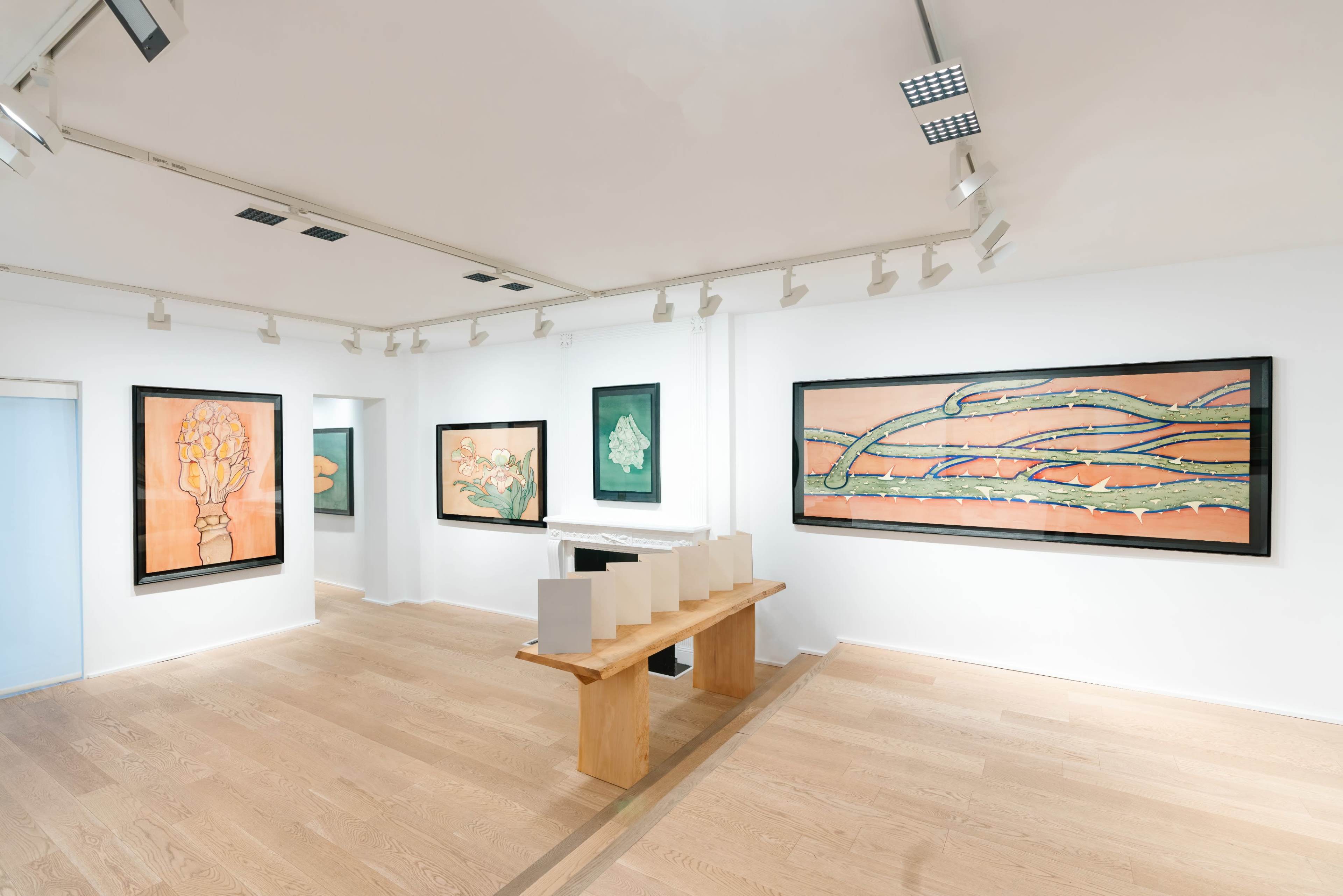 A modern art gallery features several large framed paintings on white walls, with a wooden table displaying white objects in the center of the room.
