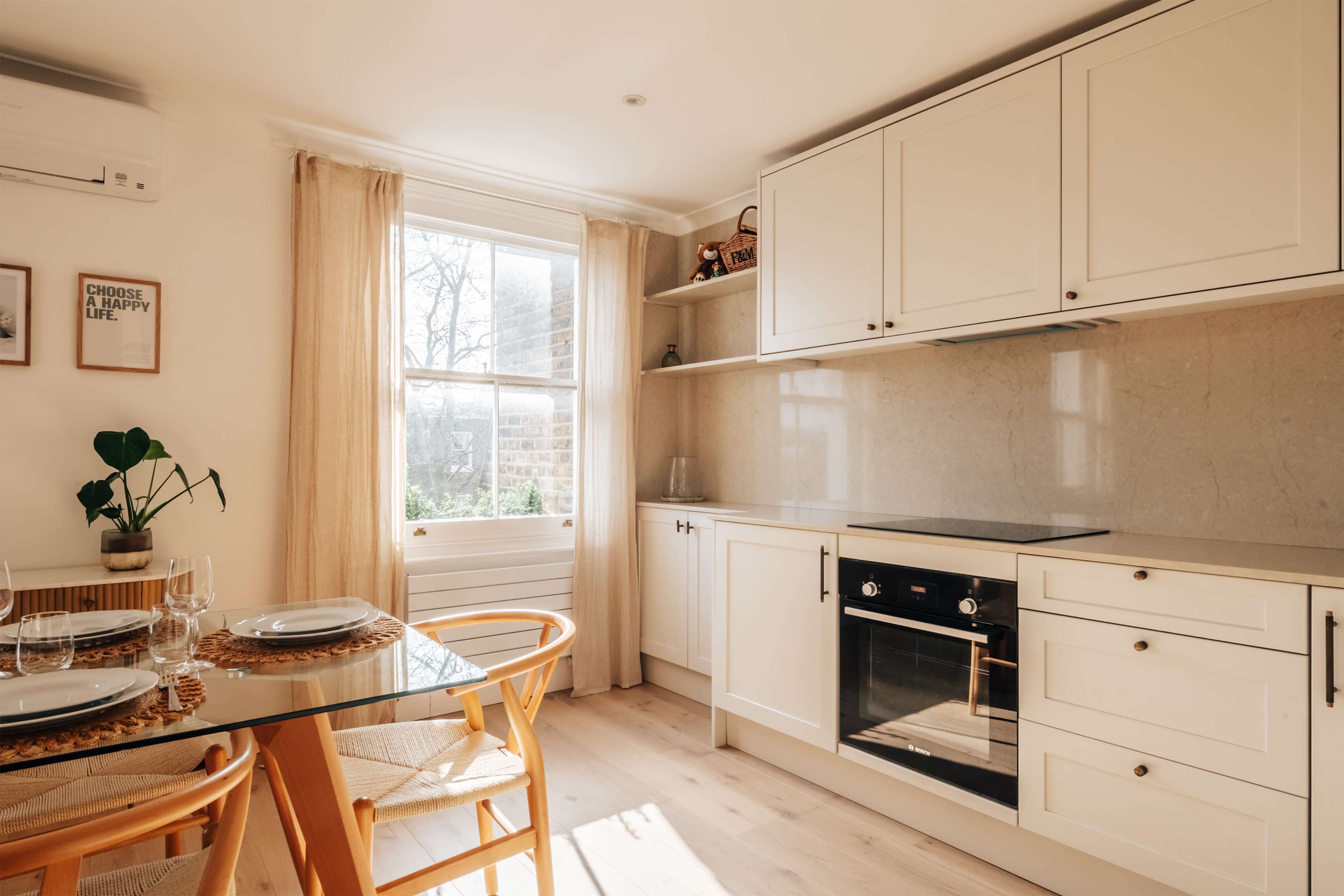 The image shows a bright kitchen with white cabinetry, a glass dining table, and a window allowing natural light to enter.