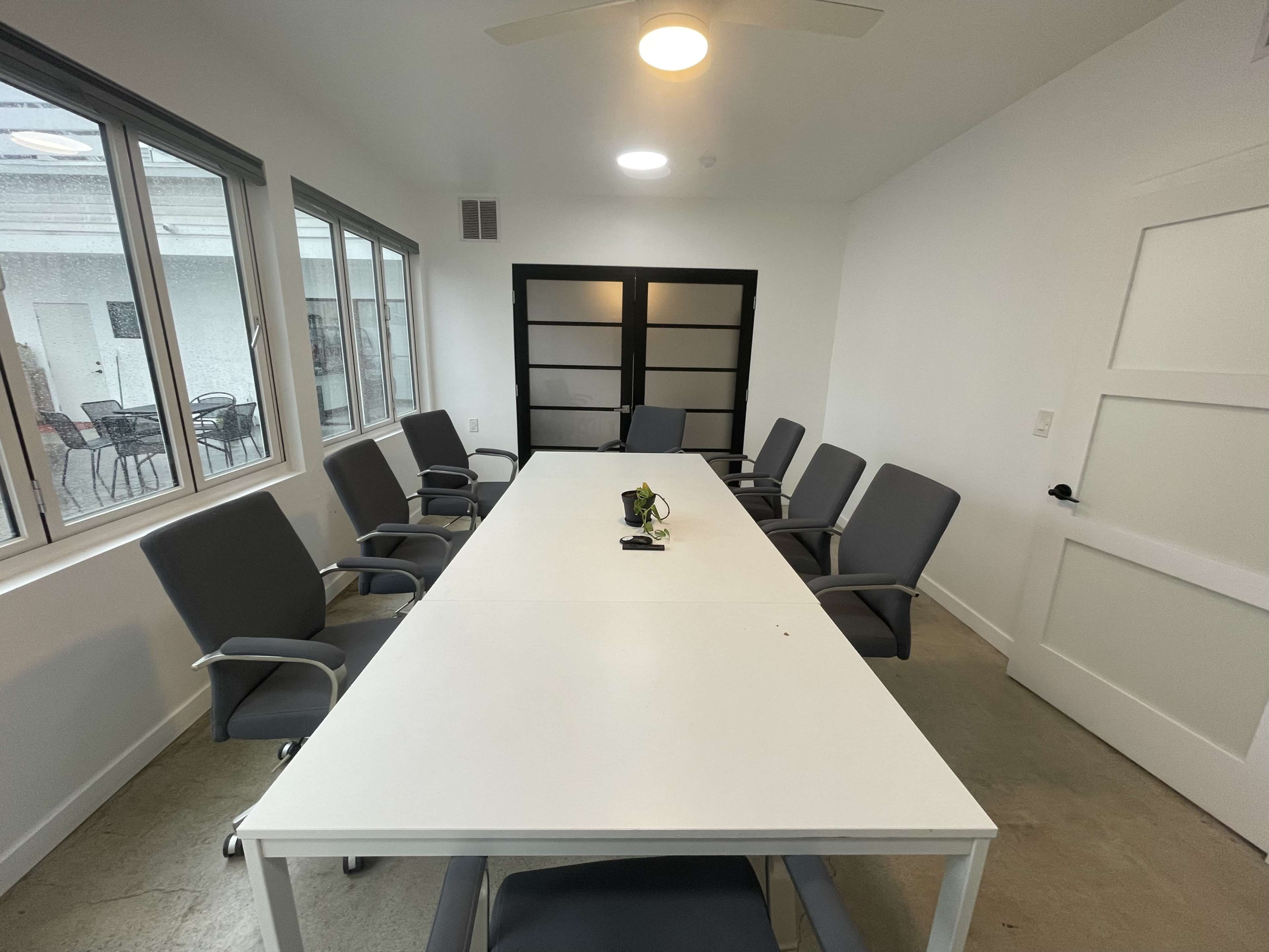 A bright, modern conference room features a large rectangular table surrounded by eight gray chairs and several windows.
