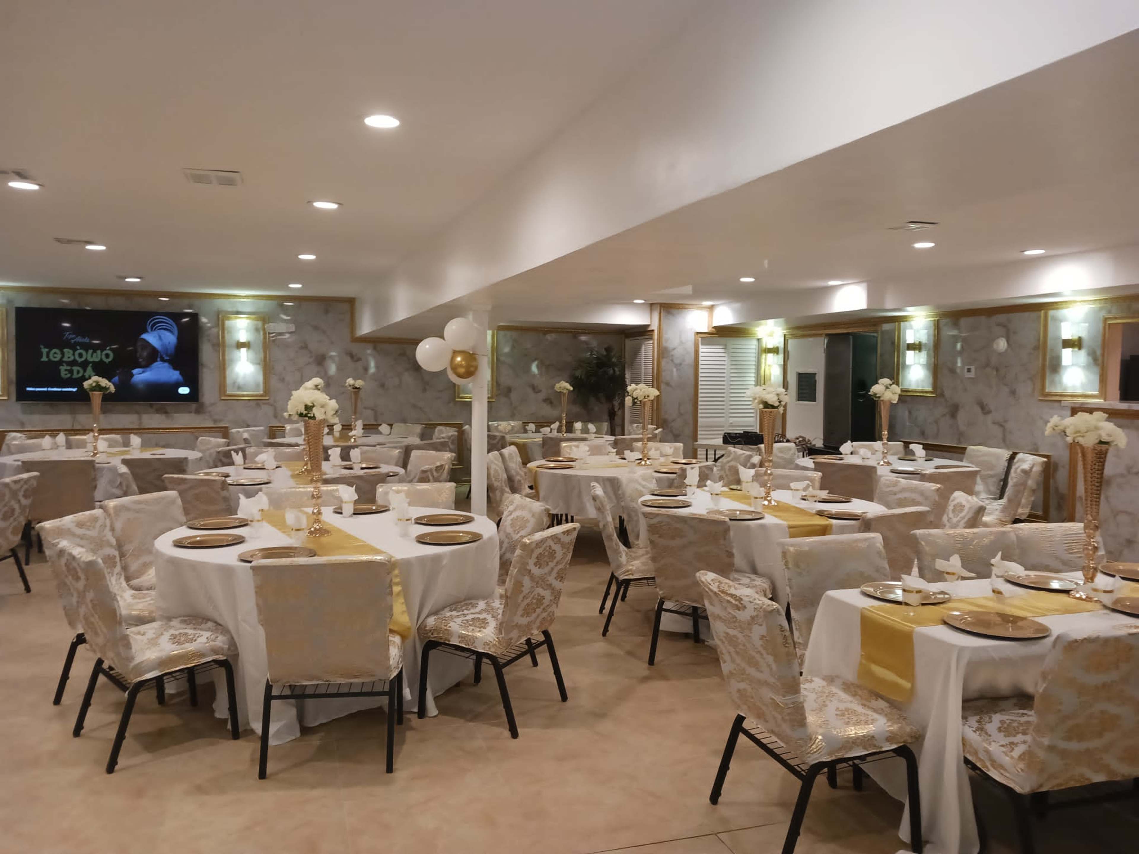 A banquet hall is arranged with neatly set tables featuring white tablecloths and gold accents, surrounded by elegant floral centerpieces.