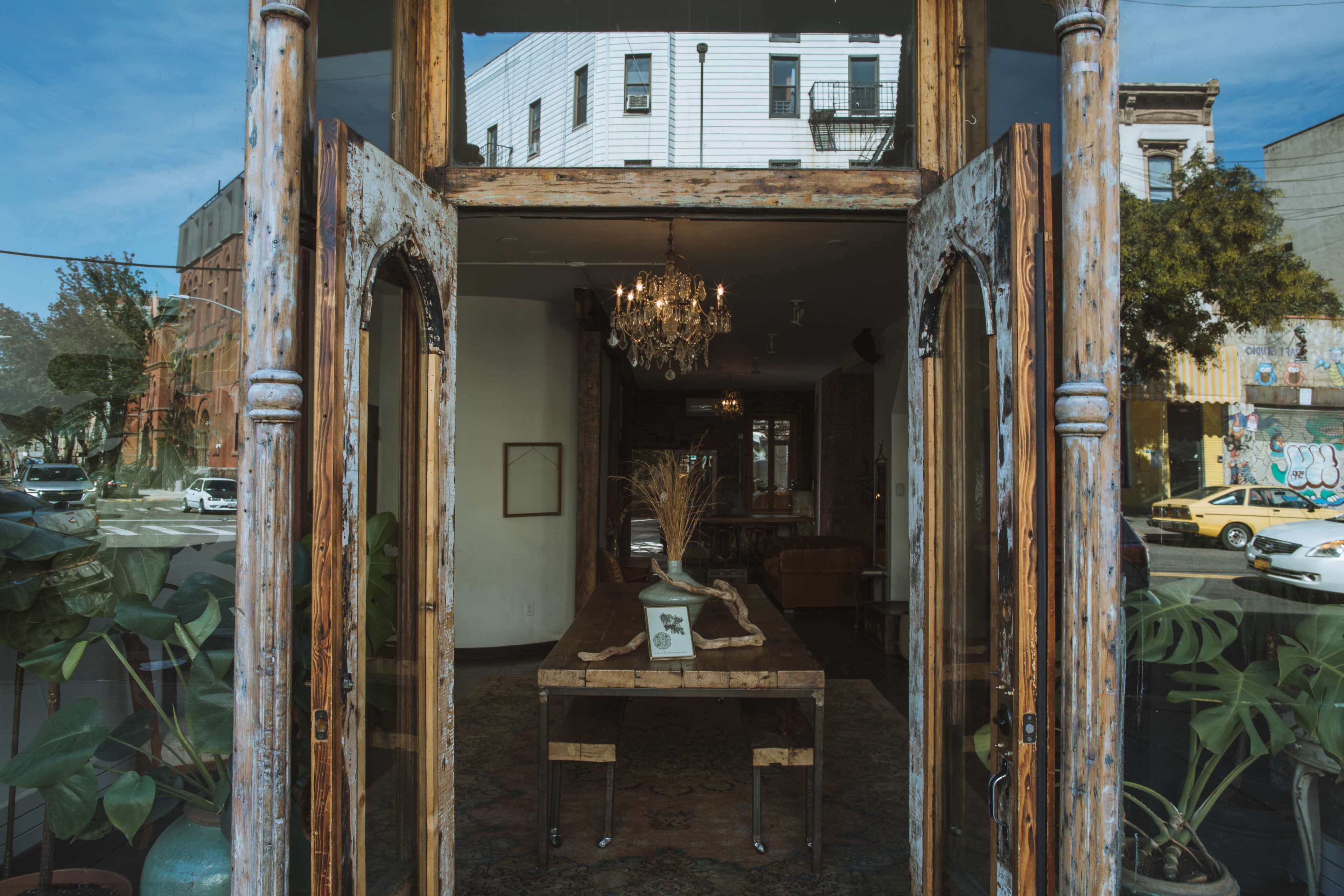 A vintage double door opens to reveal a well-lit interior with a decorative table and a chandelier, framed by large windows that showcase views of the street outside.