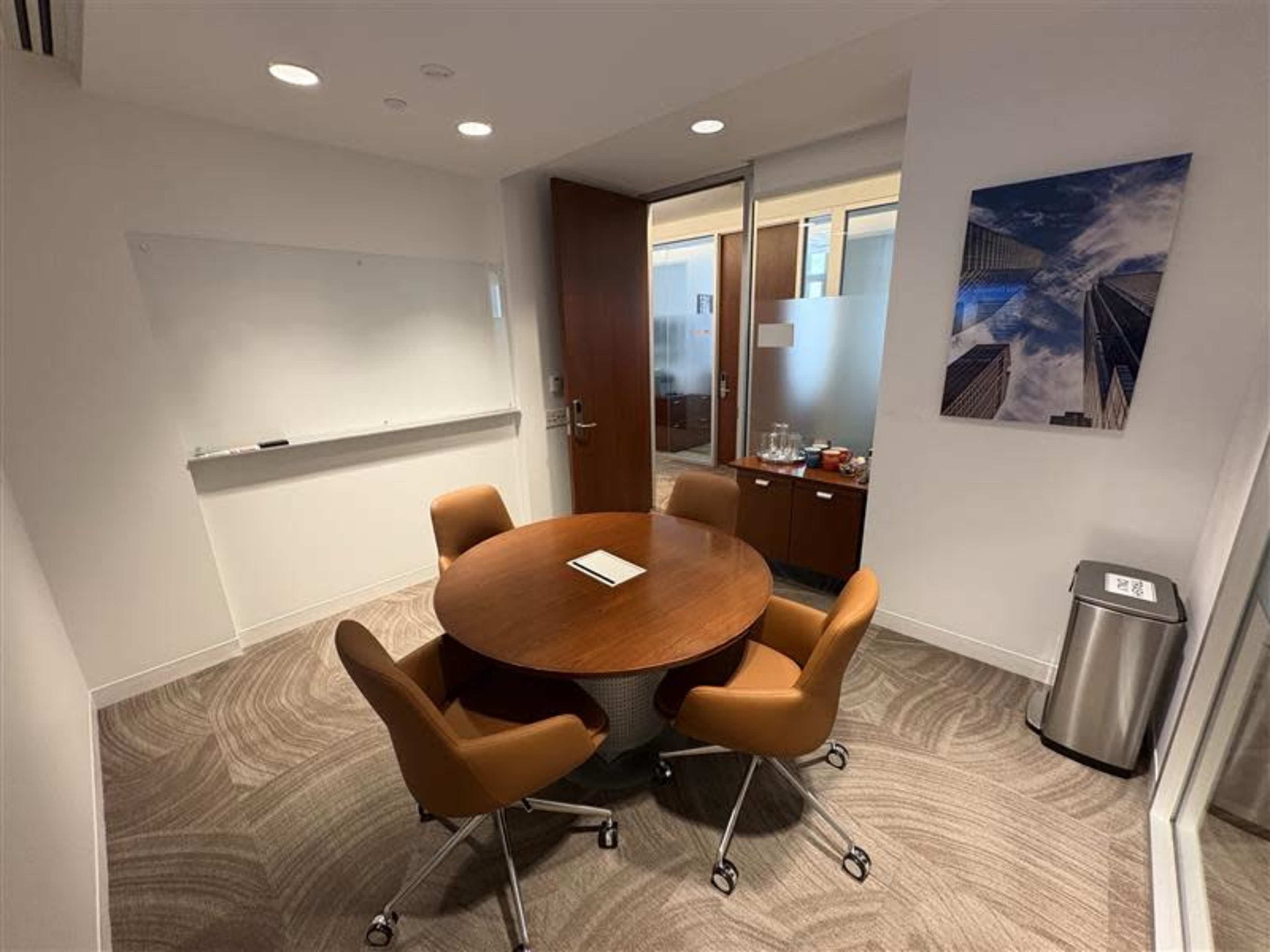 The image shows a small conference room with a circular wooden table surrounded by four chairs, a whiteboard on one wall, and a window offering a view of another office space.