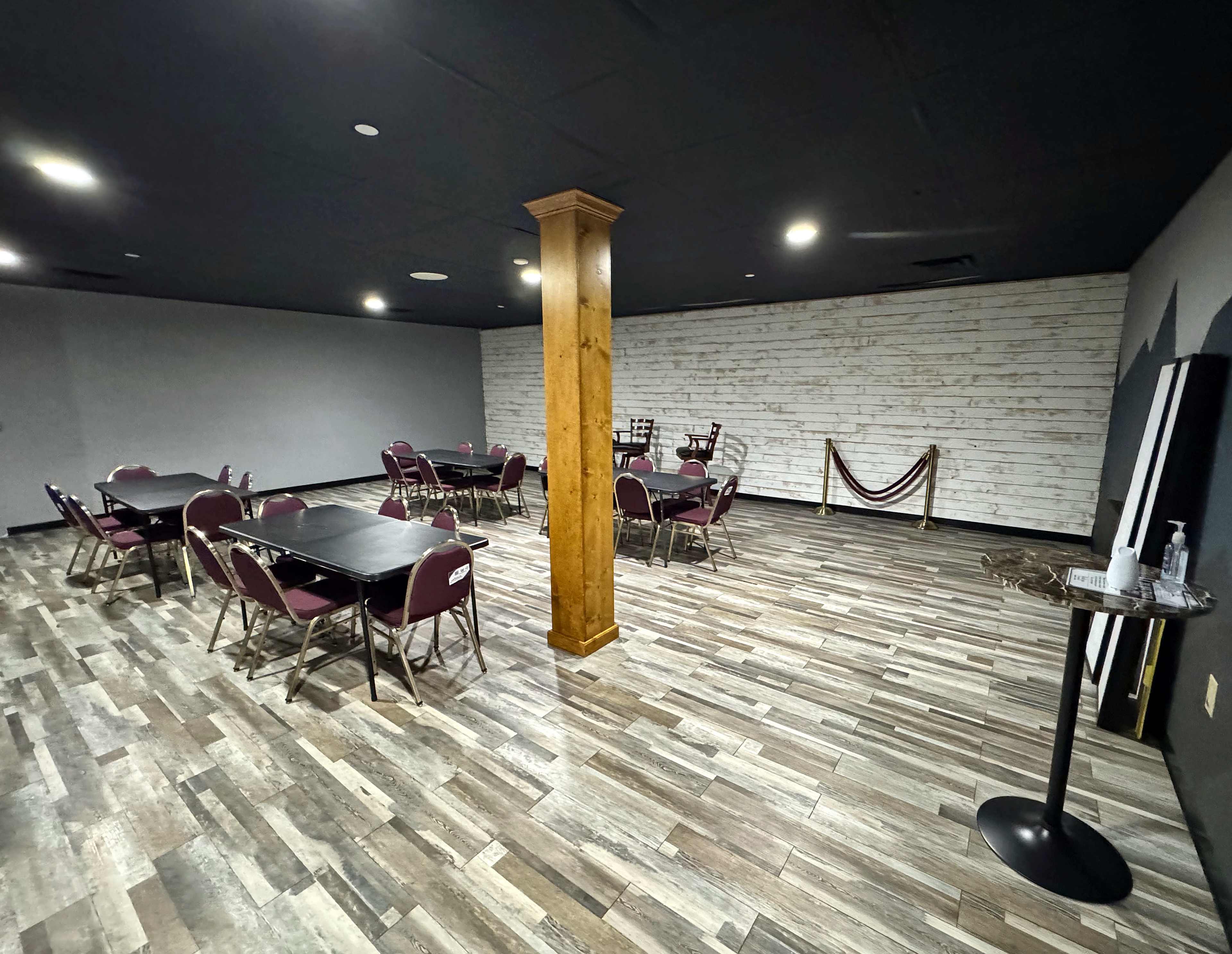 The image shows a spacious room with a wooden post in the center, featuring several tables and chairs arranged around the space, along with a small stand for refreshments.