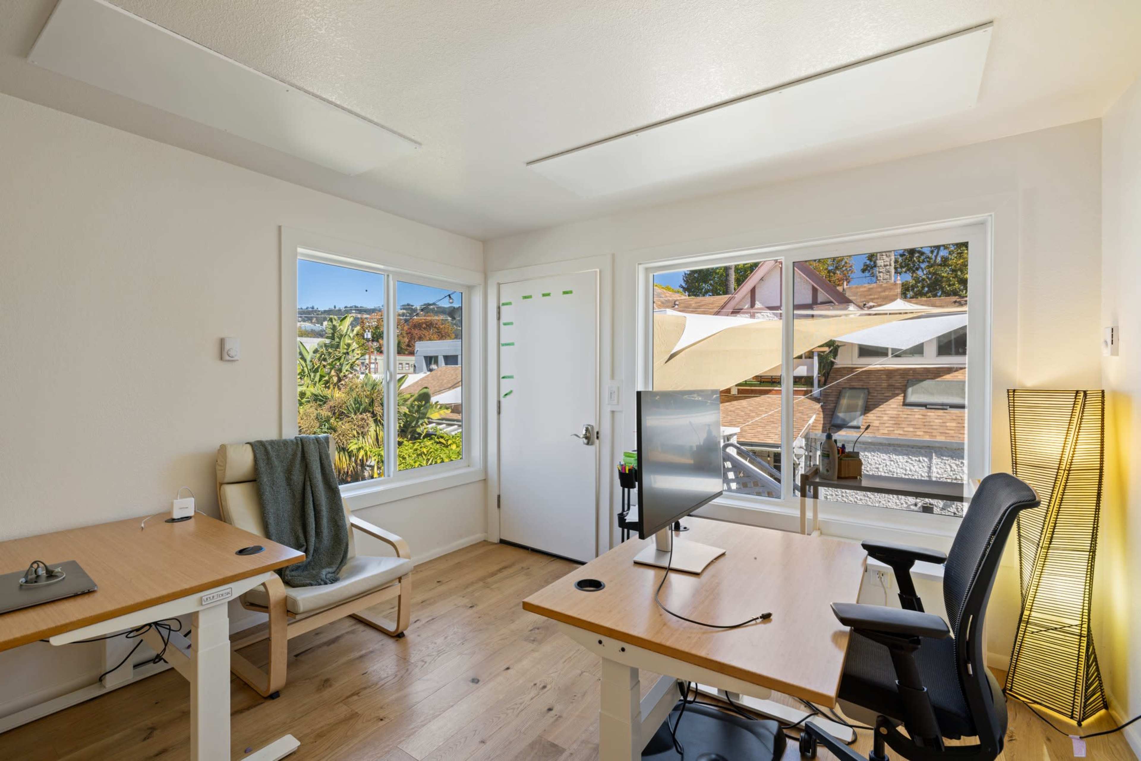 The image shows a bright office space with two desks, a chair, a door, and large windows that allow natural light to enter.