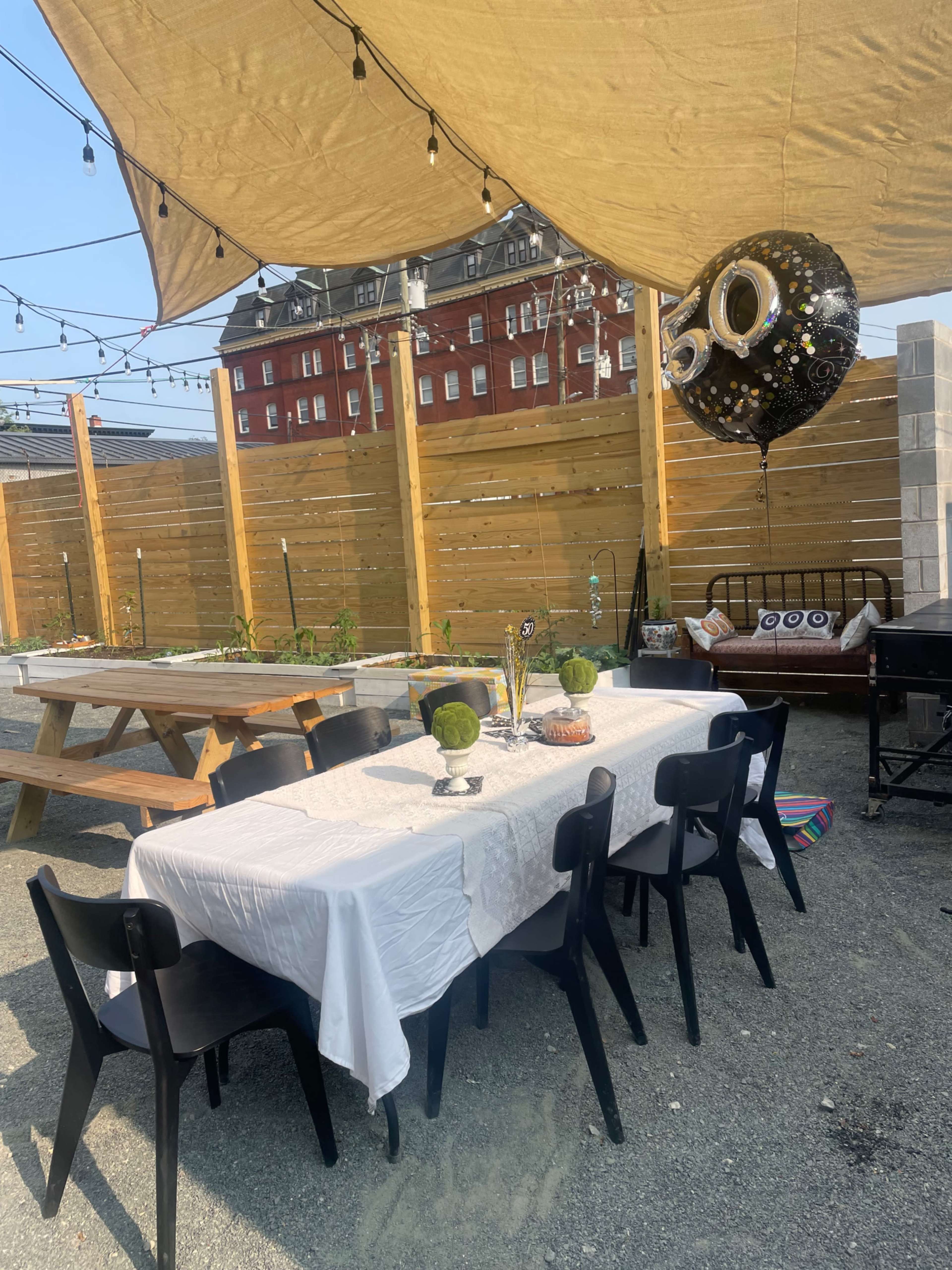 A large outdoor table is set underneath a canopy, surrounded by black chairs, with a festive "50" balloon and a wooden fence in the background.