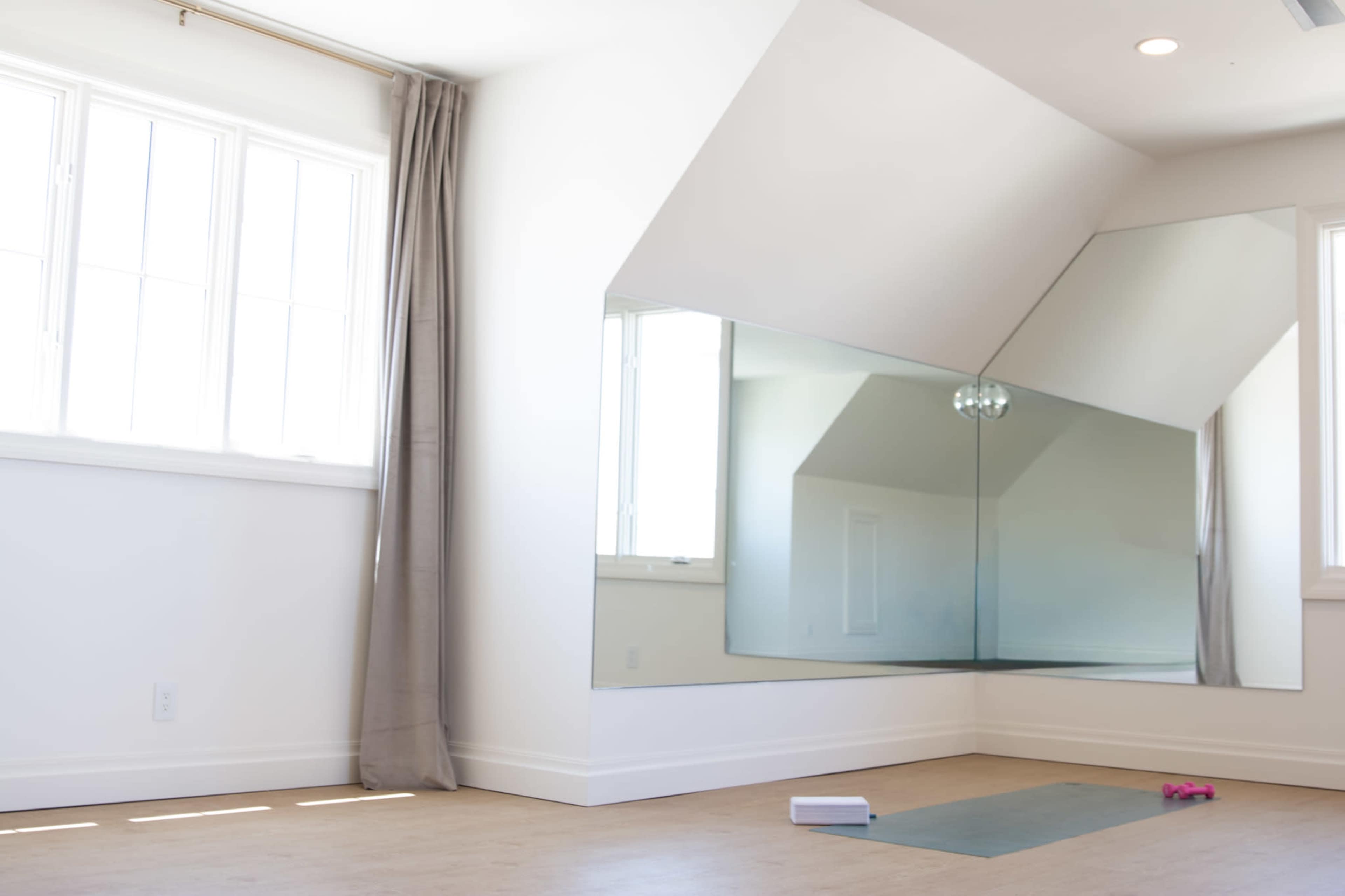 A spacious, well-lit room features a large mirrored wall, a yoga mat on the floor, and a set of pink weights nearby.