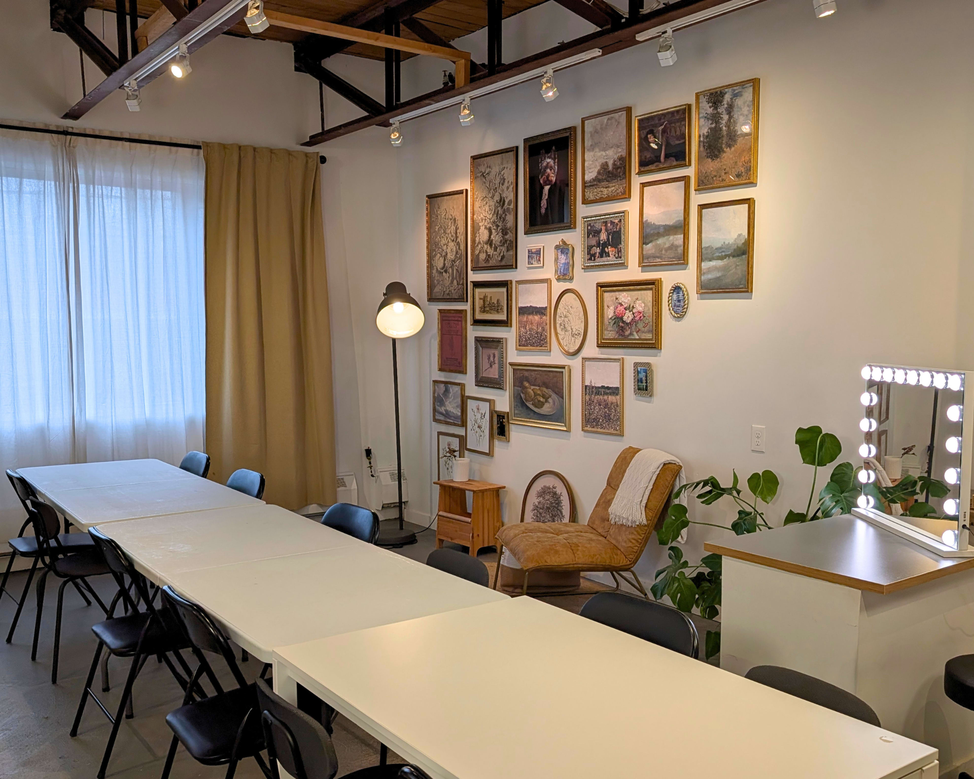 A well-lit room features a long white table surrounded by black chairs, with a gallery wall of framed pictures and a cozy chair near a lamp.