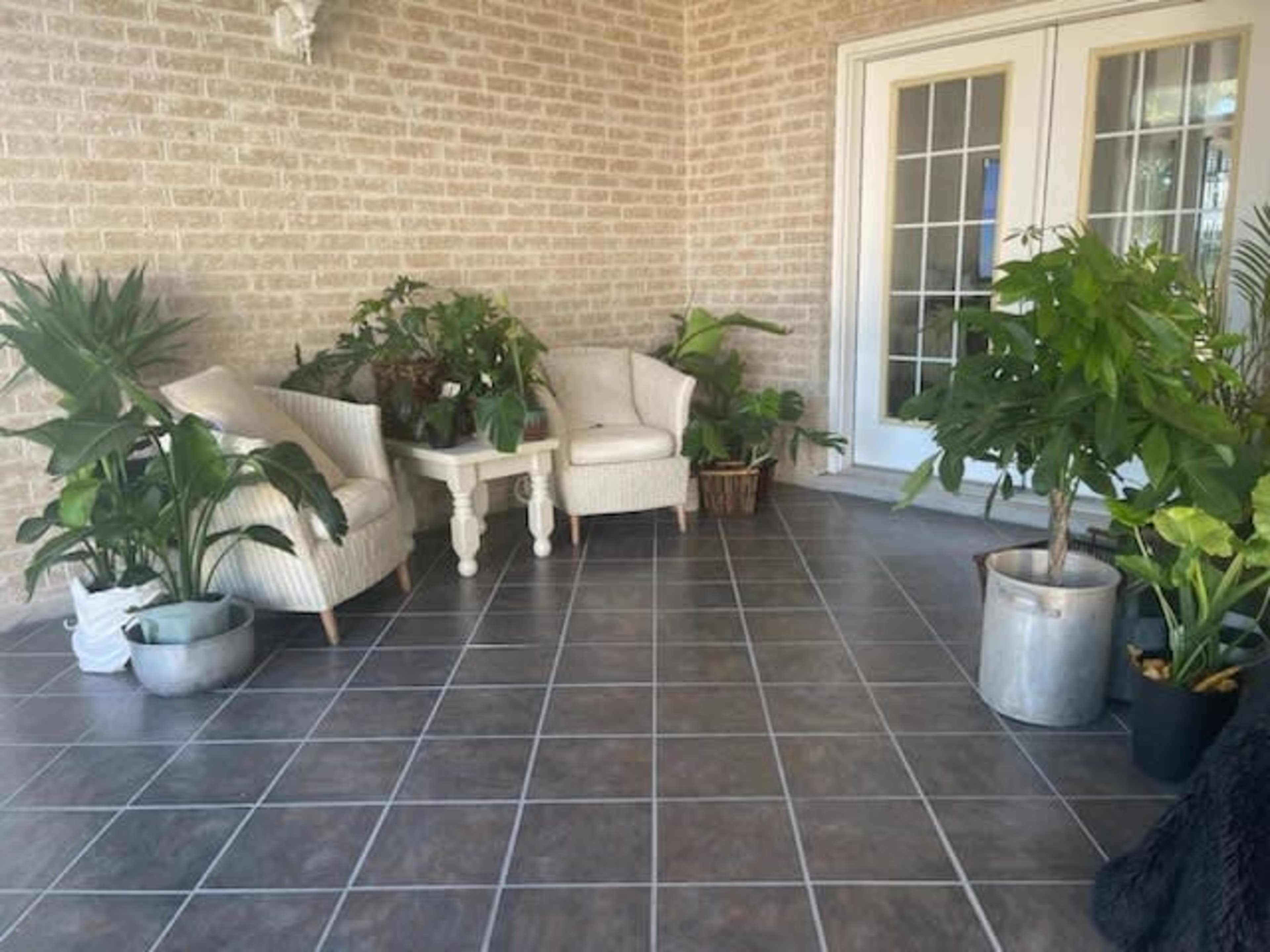 A patio area features two white armchairs, a small table, and various potted plants on a tiled floor.