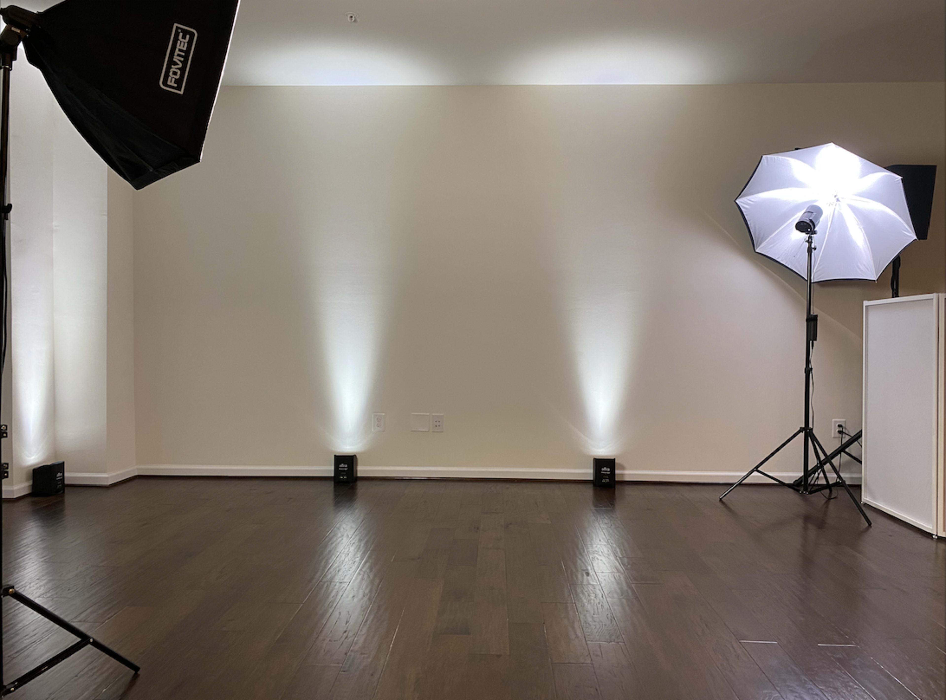 A photography studio setting with two light sources, one softbox and one strobe light, positioned in a room with hardwood flooring and a plain wall.