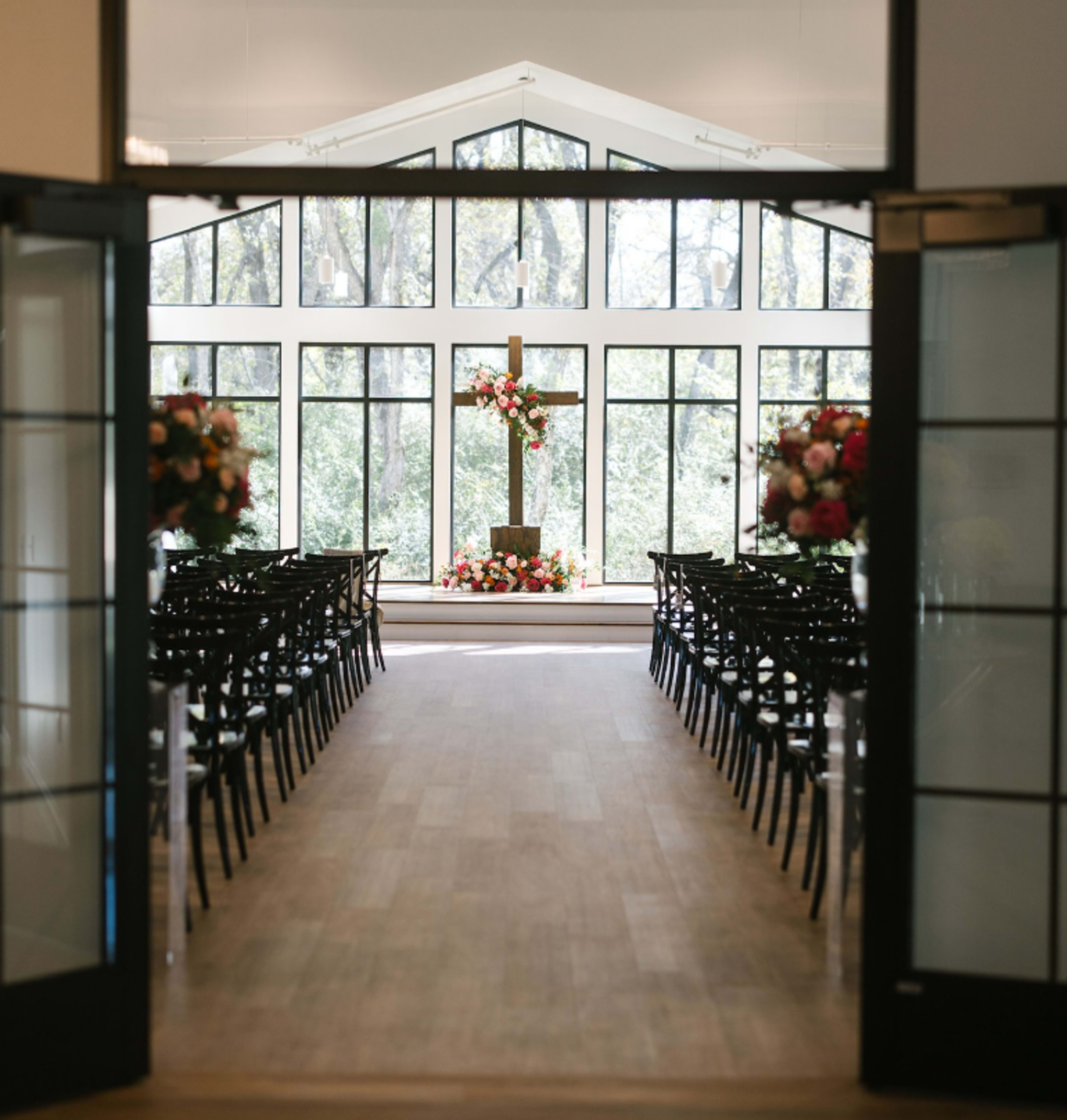 A wedding ceremony space features rows of black chairs, a floral-adorned cross at the front, and large windows showcasing a natural backdrop.