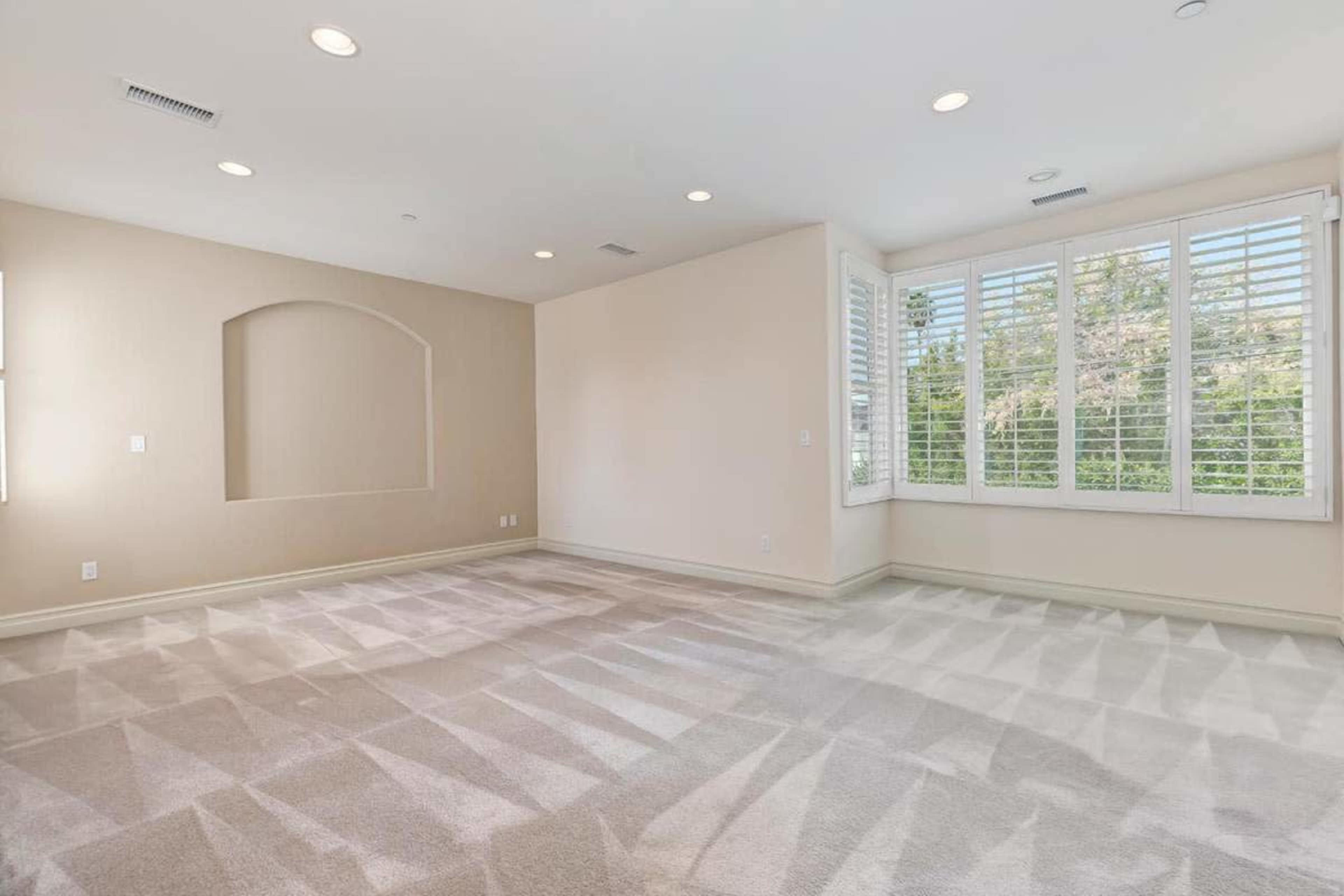 The image shows an empty room with light-colored walls, carpeted flooring, and large windows with shuttered blinds.